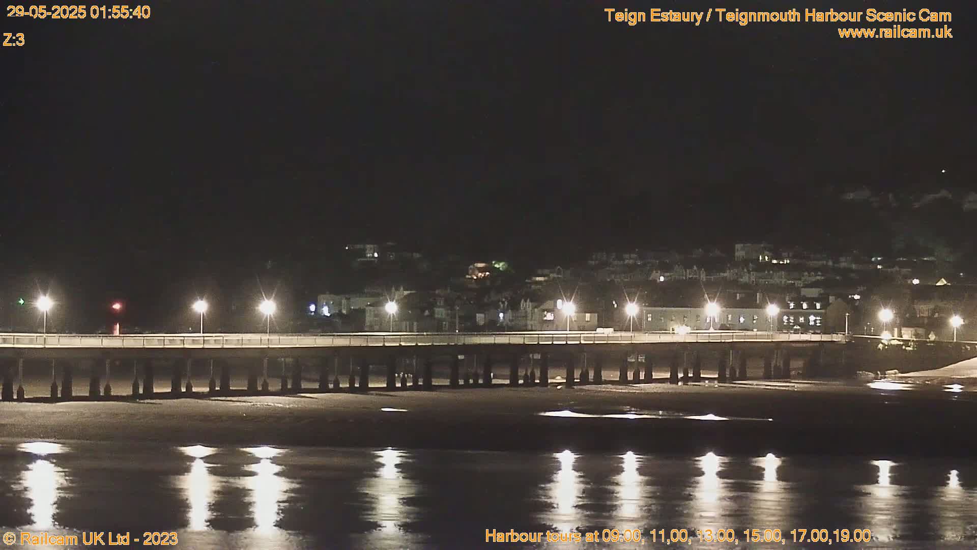 A long pier with lights extends over a dark, calm body of water at night, with a town visible in the background.