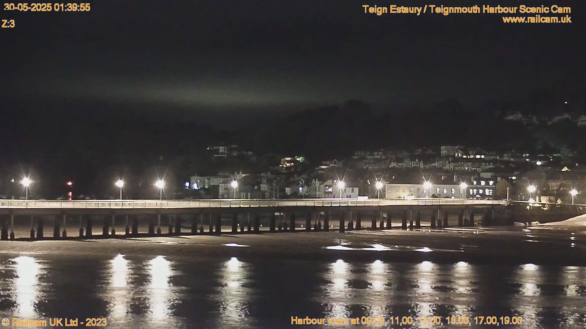 A long pier is illuminated at night over calm, dark water, with a town visible in the background under a dark sky.