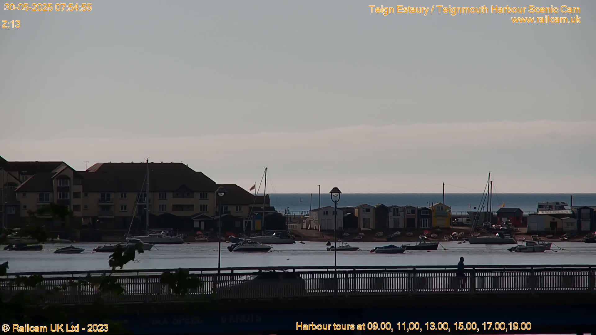 A calm, overcast day at a harbor, showing boats moored near a building and a bridge with a person walking on it.