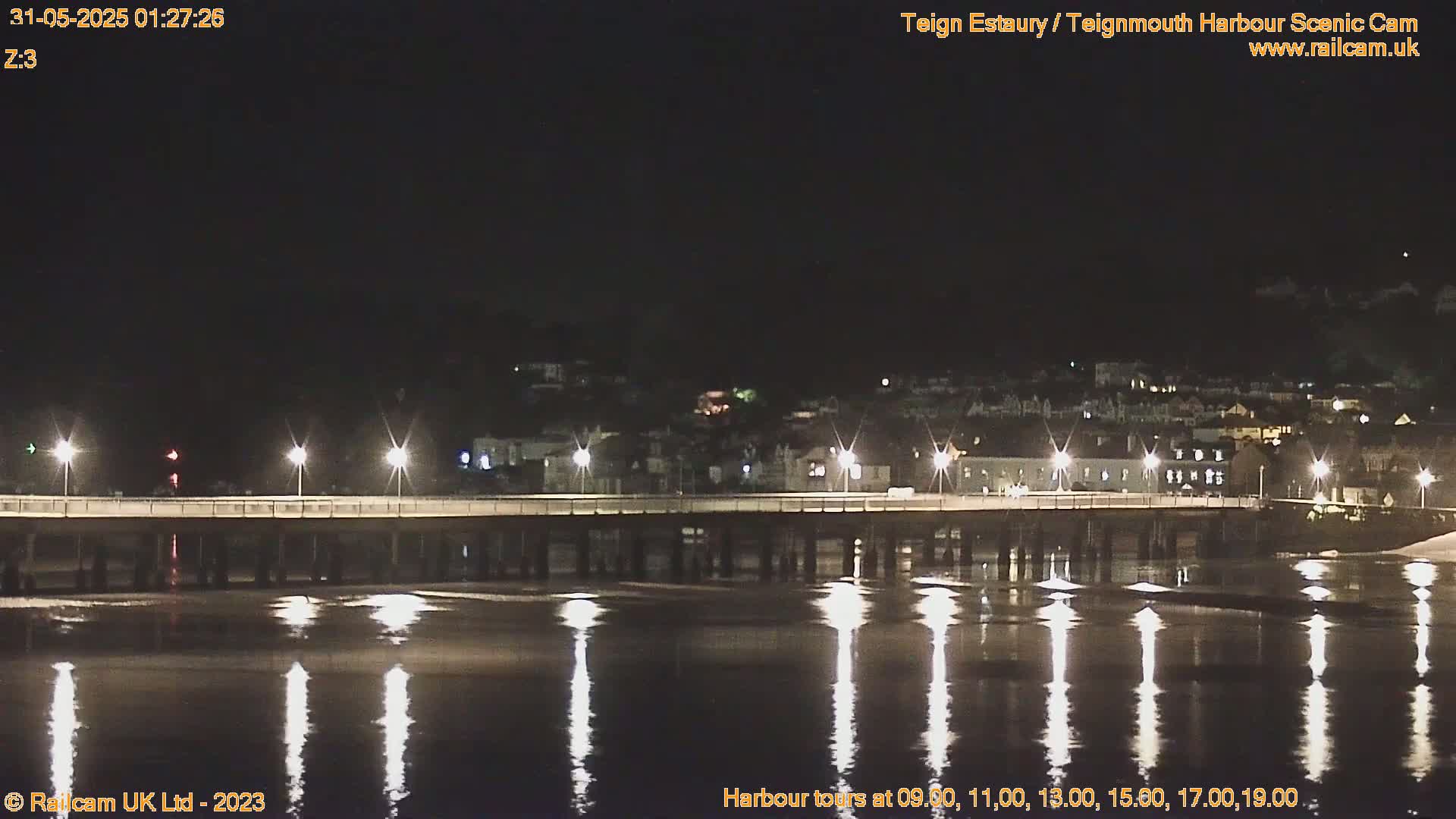 A long, dark pier is illuminated by lights at night, reflecting in calm water under a clear, dark sky.
