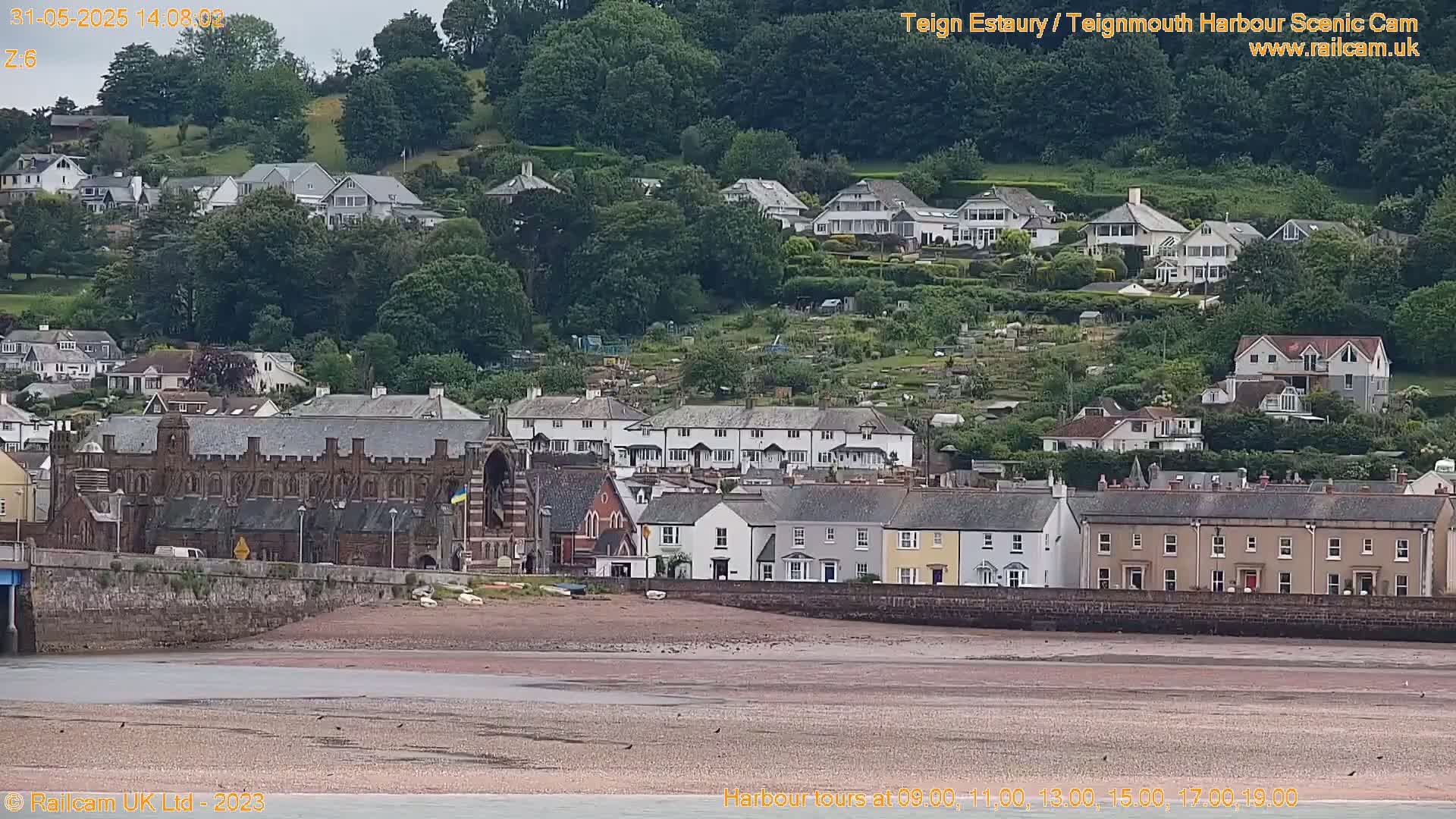 A coastal town with numerous houses and buildings sits on a hillside overlooking a wide, flat expanse of muddy beach under a mostly cloudy sky.