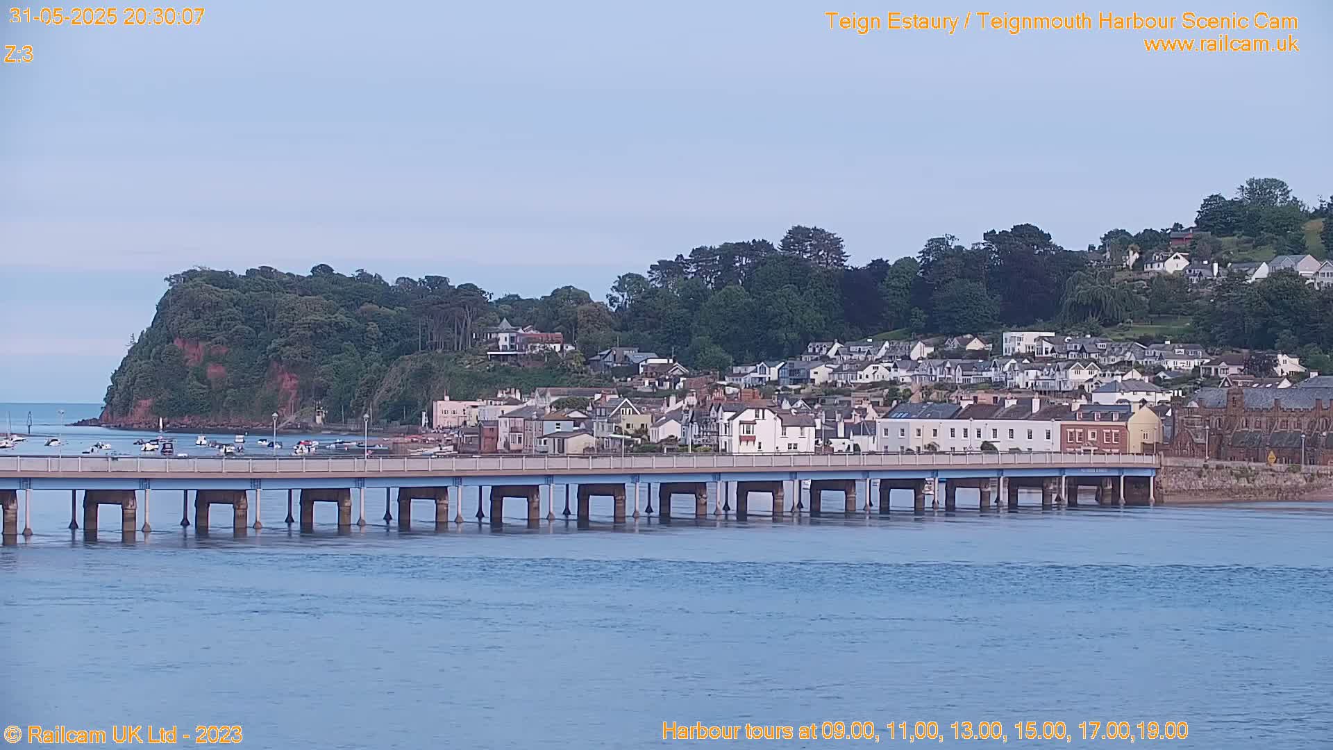 A long bridge extends over calm water toward a town nestled among green hills under a clear, twilight sky.