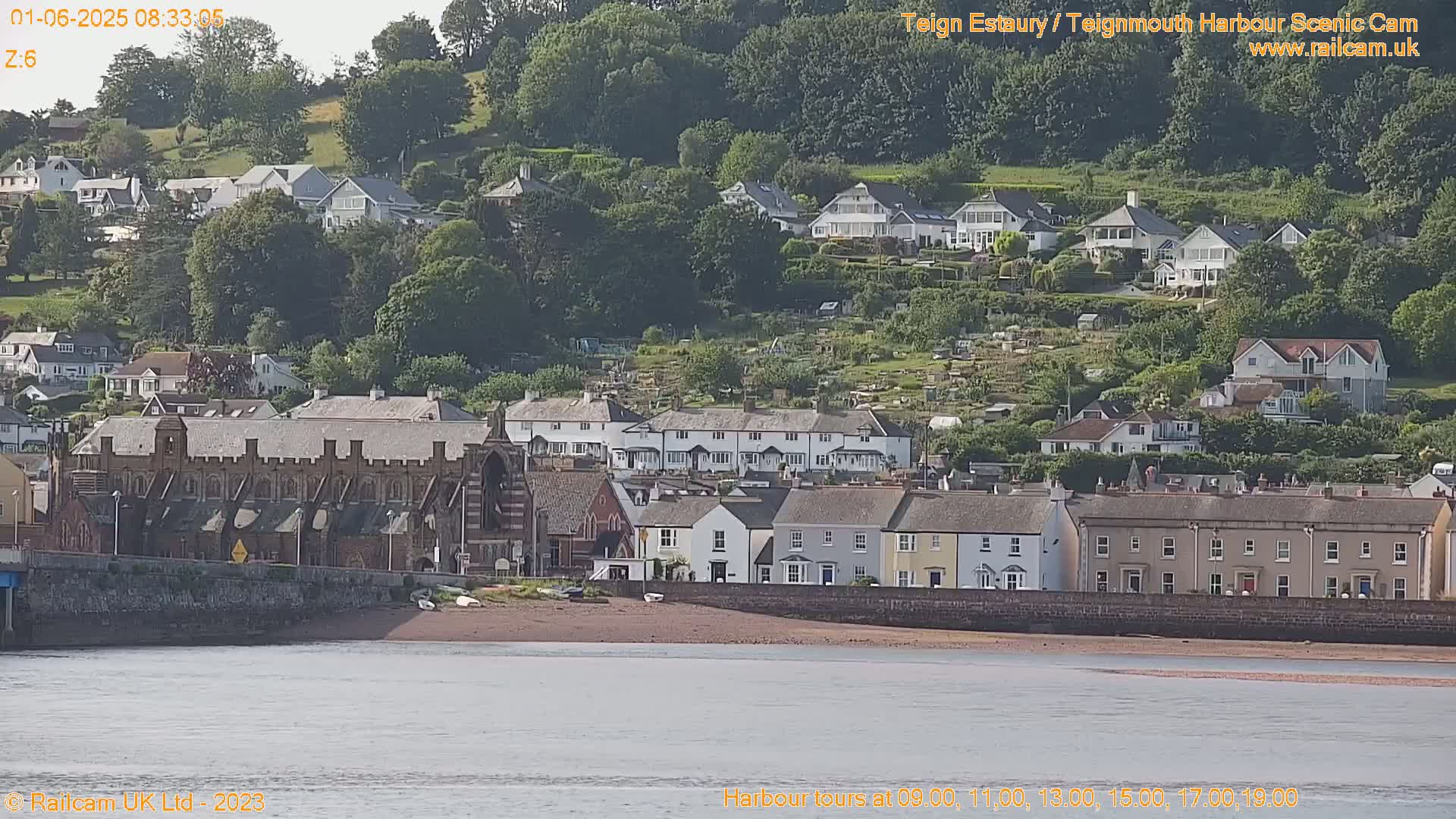 A brick building sits on a waterfront next to a row of houses and a hillside of more houses and green space under a partly sunny sky.
