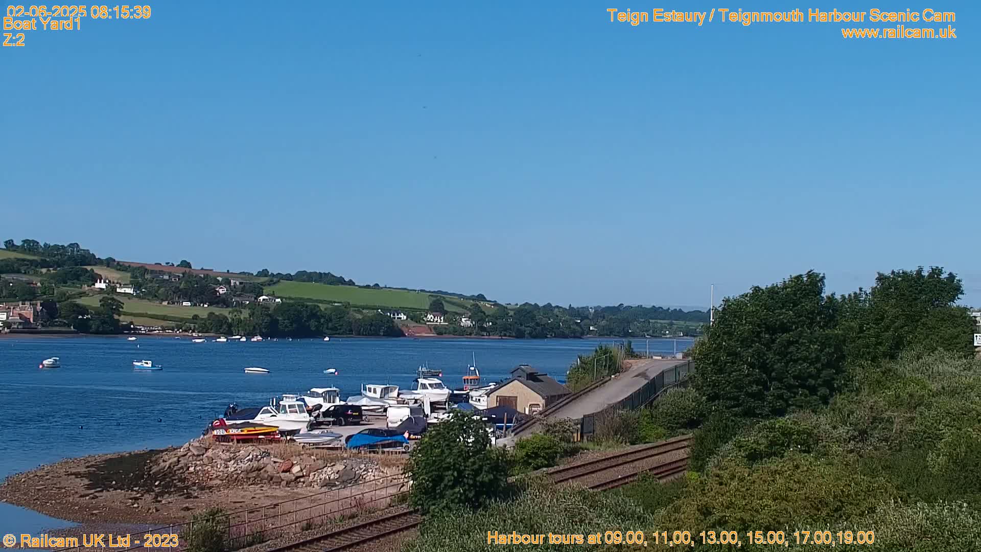 A sunny day shows a calm river with several boats moored at a boat yard next to train tracks, all situated near a verdant hillside.