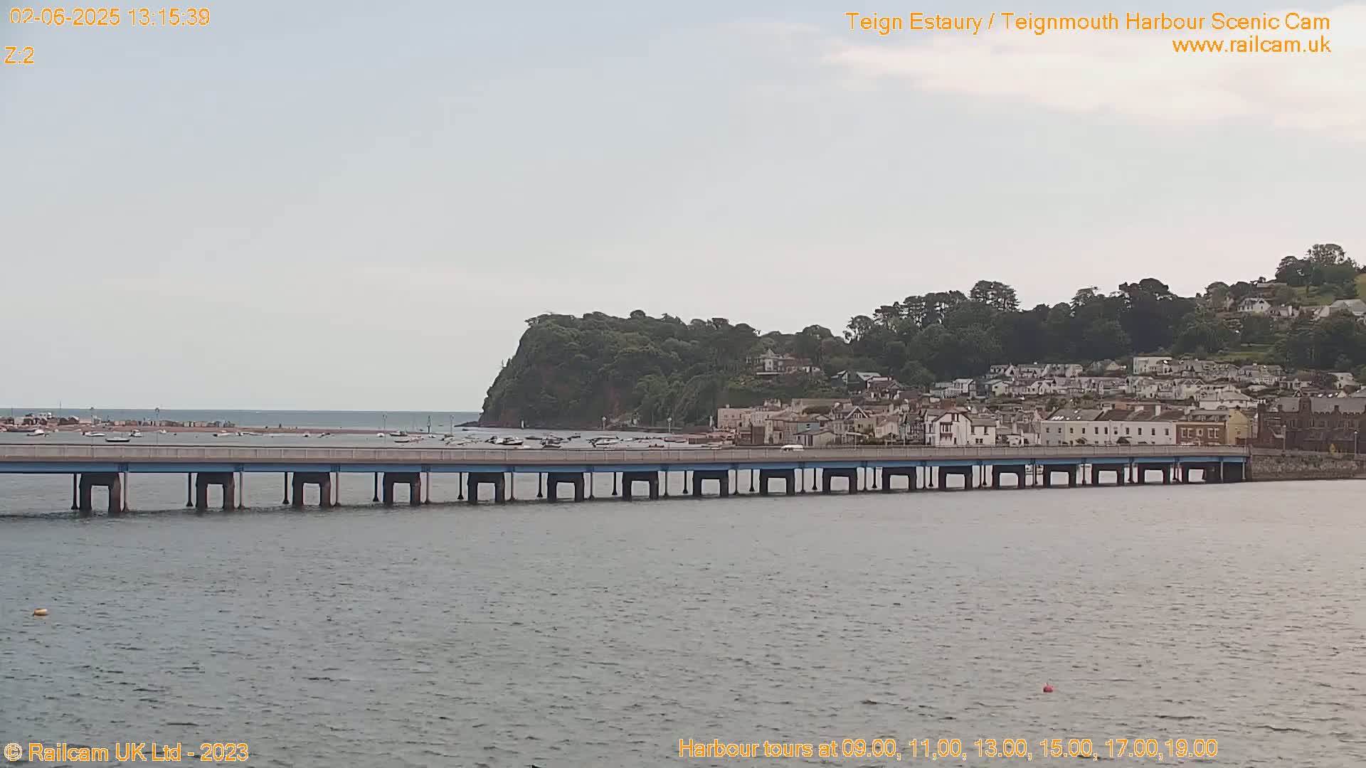 A long bridge spans a calm body of water, with a town nestled against a hillside in the background under an overcast sky.