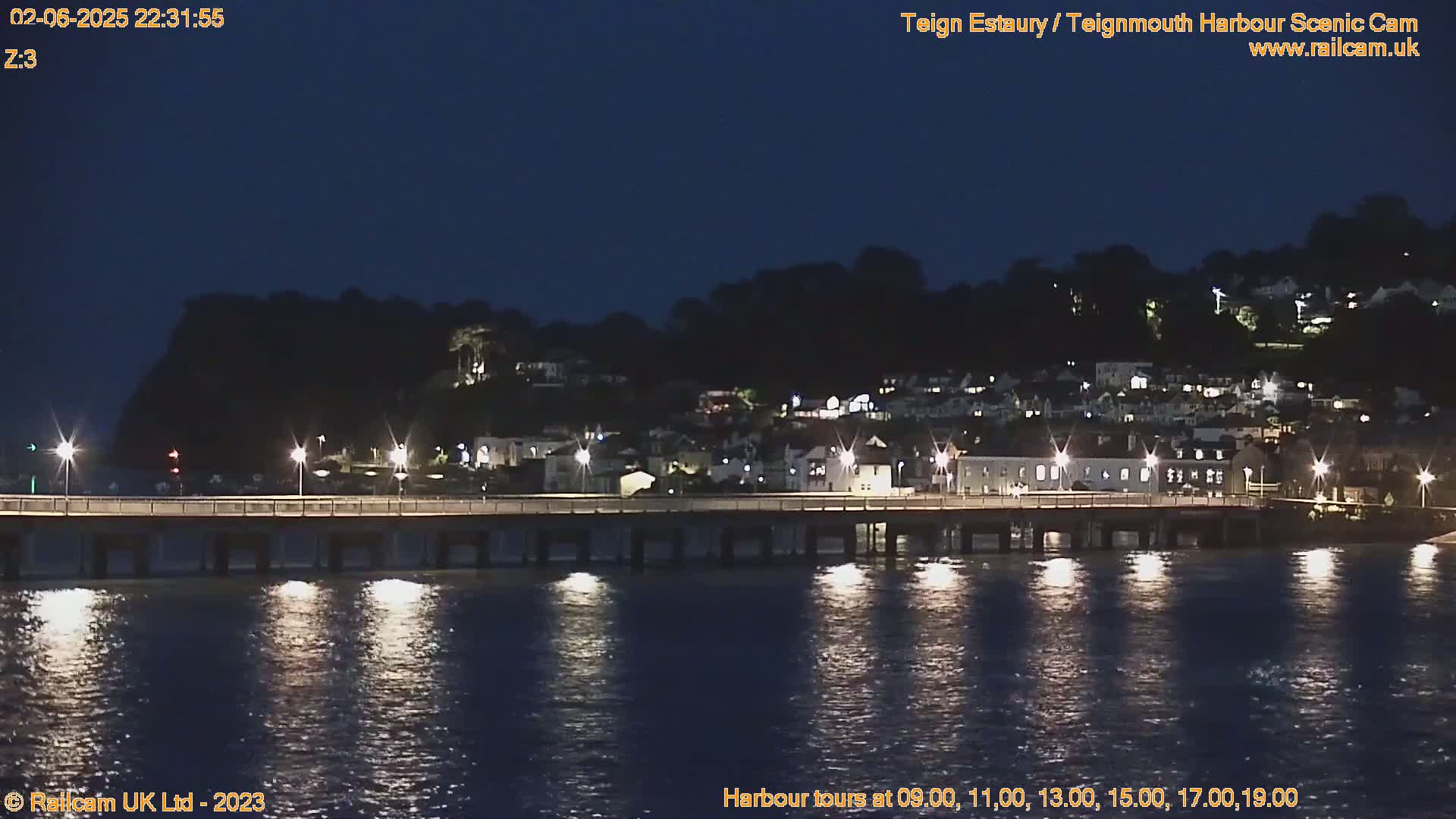 A nighttime view of a lit-up harbor town across a calm body of water, with a bridge connecting the town to the opposite shore.