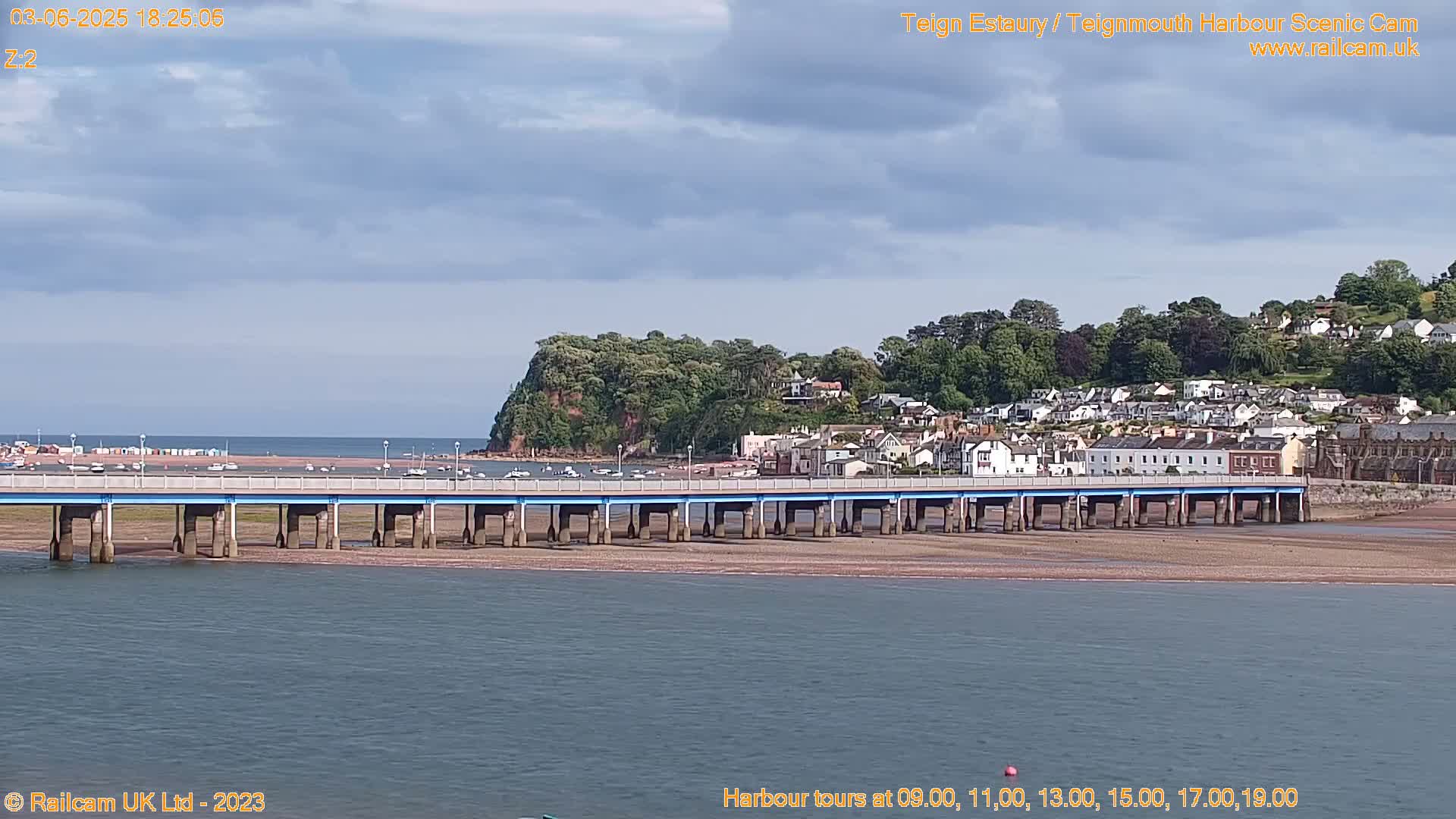 A long bridge stretches across a calm body of water towards a town nestled on a hillside under a partly cloudy sky.