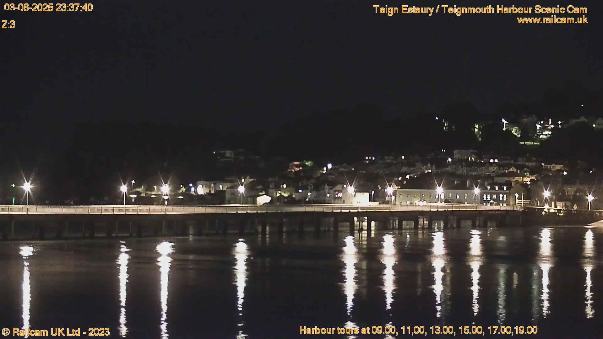 A long pier illuminated at night stretches across calm, reflective water toward a dark hillside town.