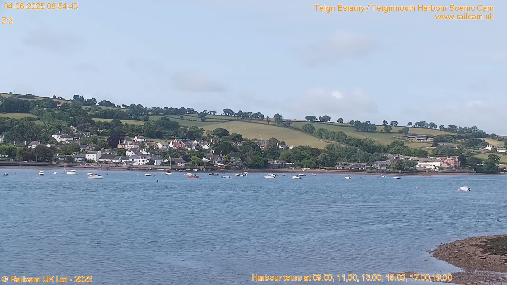 A calm body of water shows several small boats, with a town nestled along a hillside in the background under a mostly clear sky.