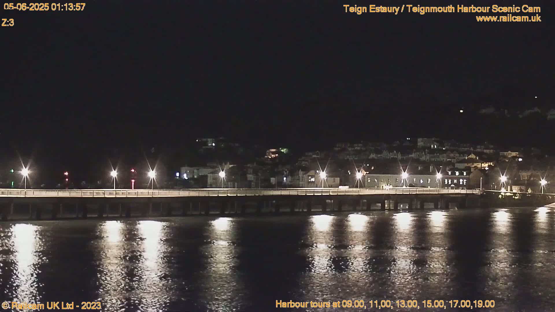 A long pier, illuminated by lights, extends over dark water at night, with a town visible in the background.