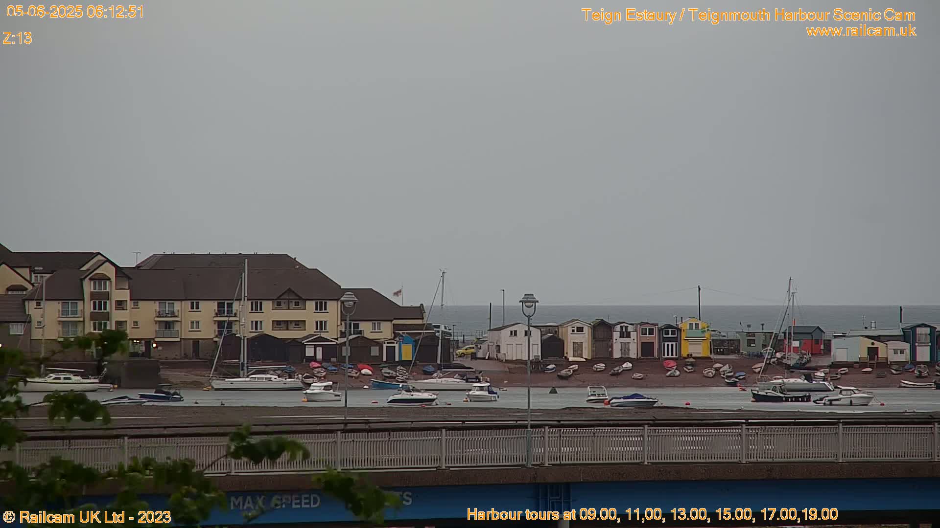 A harbor scene on an overcast day shows numerous boats moored along a shoreline with buildings and a bridge in the foreground.