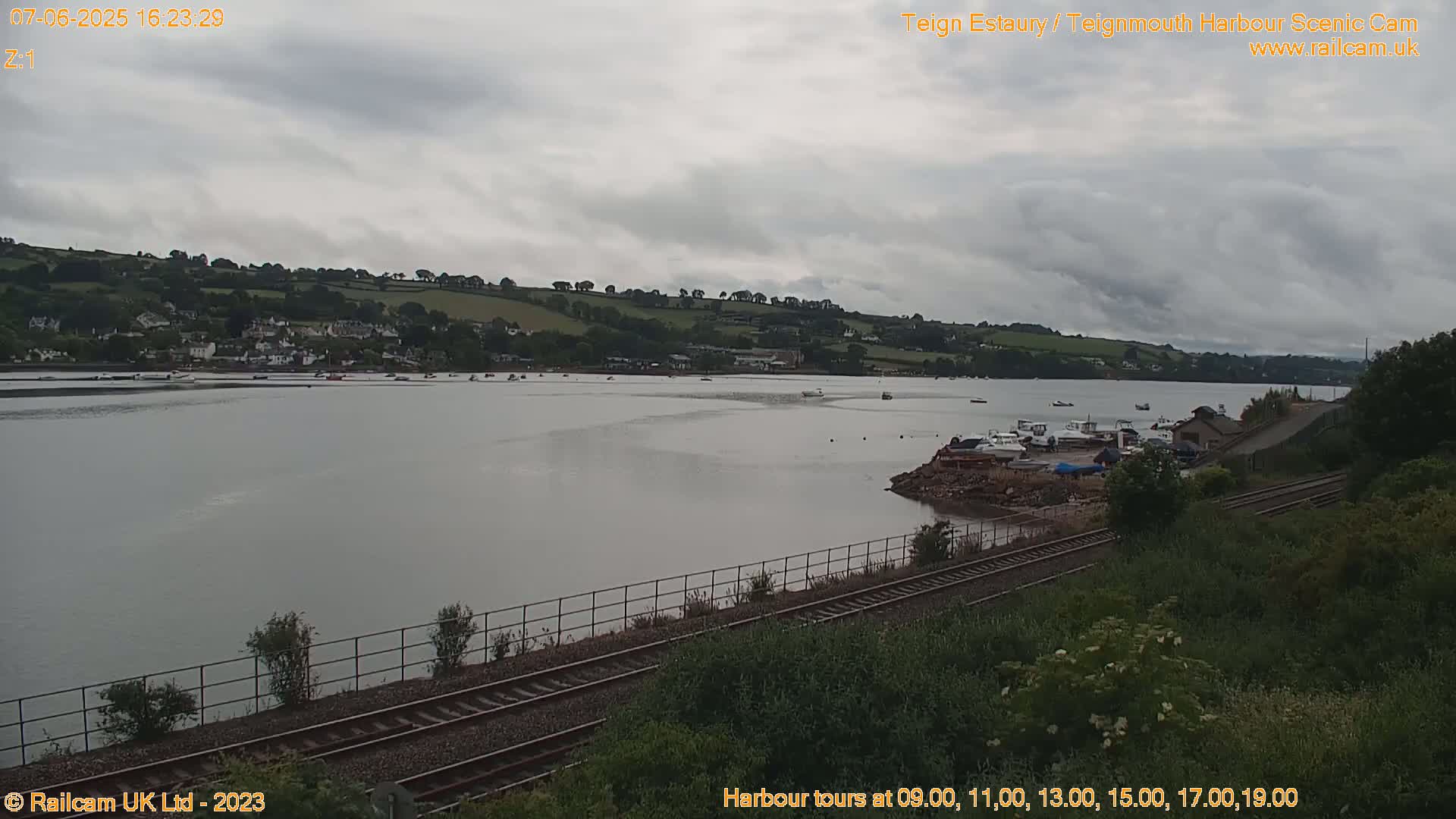 A calm body of water with several boats is viewed from a slightly elevated vantage point showing train tracks and green vegetation in the foreground under an overcast sky.