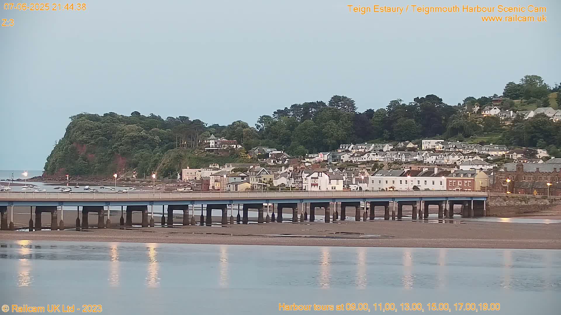 A bridge spans a calm, reflective body of water at dusk, overlooking a town nestled against a verdant hillside.
