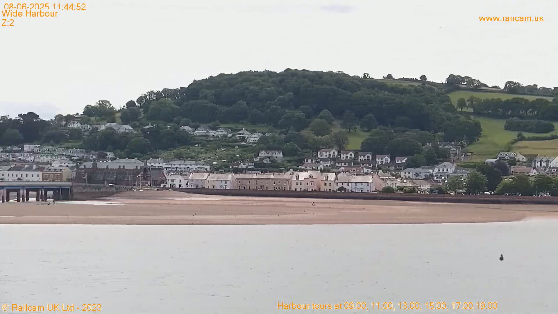 A coastal town with light-colored buildings sits beneath a verdant hill under an overcast sky, with a sandy beach and calm water in the foreground.