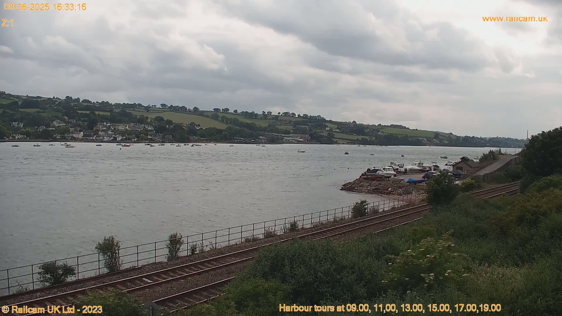 A mostly cloudy day reveals a tranquil harbor scene with numerous boats, a small town nestled against a verdant hillside, and railroad tracks running alongside the water's edge.