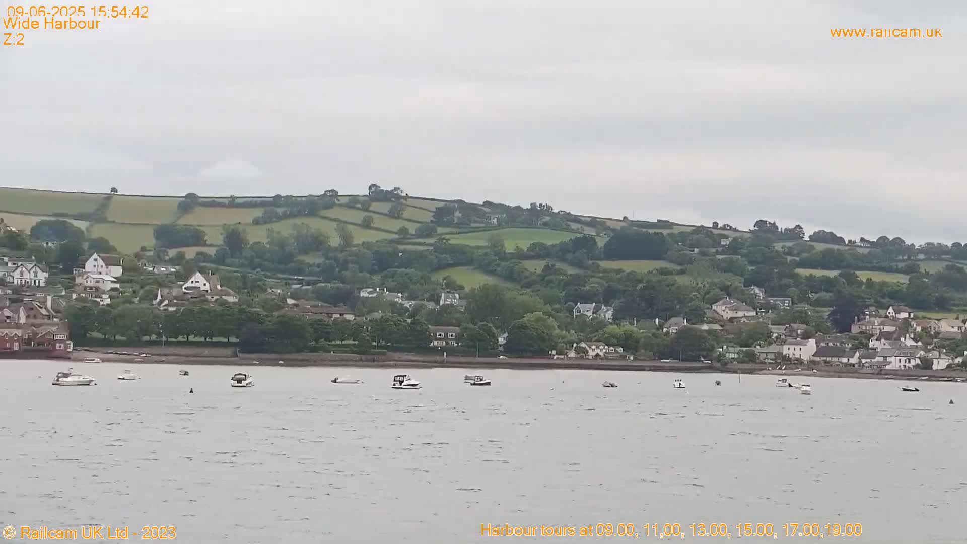 A calm body of water with several small boats is seen in front of a hilly shoreline dotted with houses and green fields under an overcast sky.