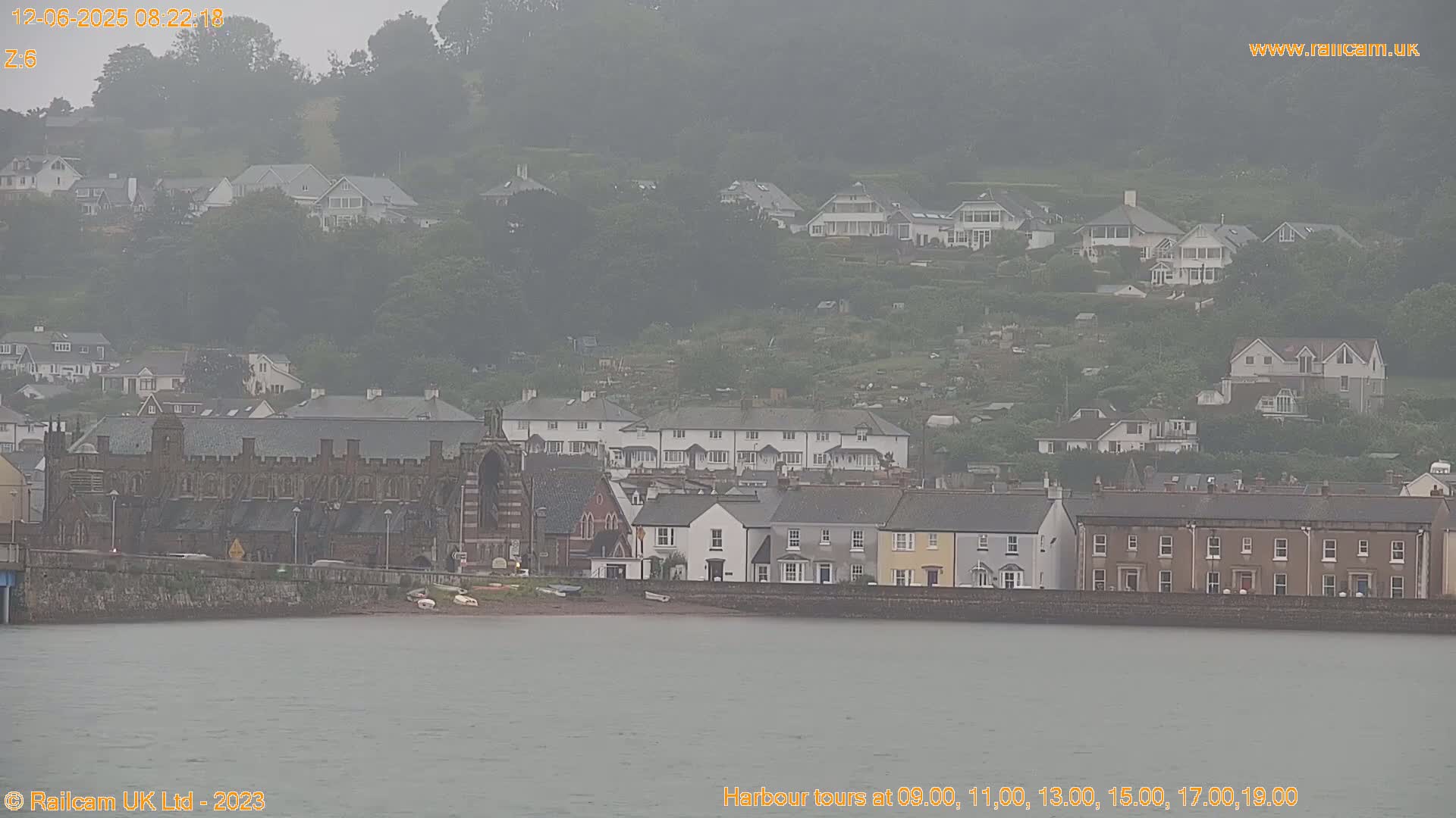 A hazy view of a coastal town on a cloudy day shows a mix of residential buildings, including several rows of terraced houses, and a large stone building near the water's edge.