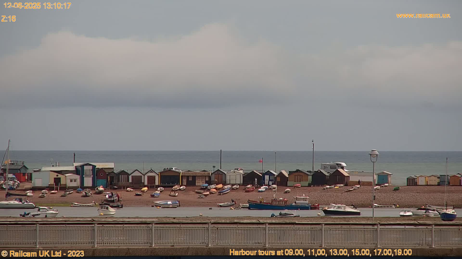 A row of colorful beach huts sits on a pebbled shore next to a calm sea under a mostly cloudy sky.