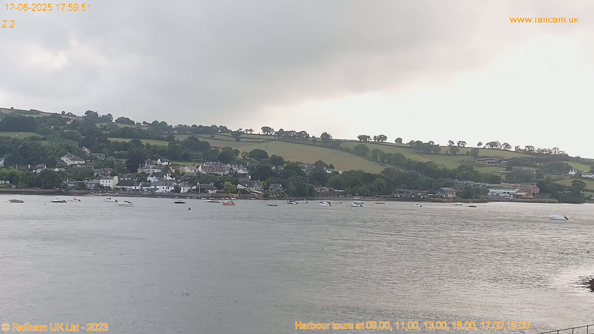 A calm body of water with several small boats is in front of a small town nestled against a gently sloping, green hill under an overcast sky.