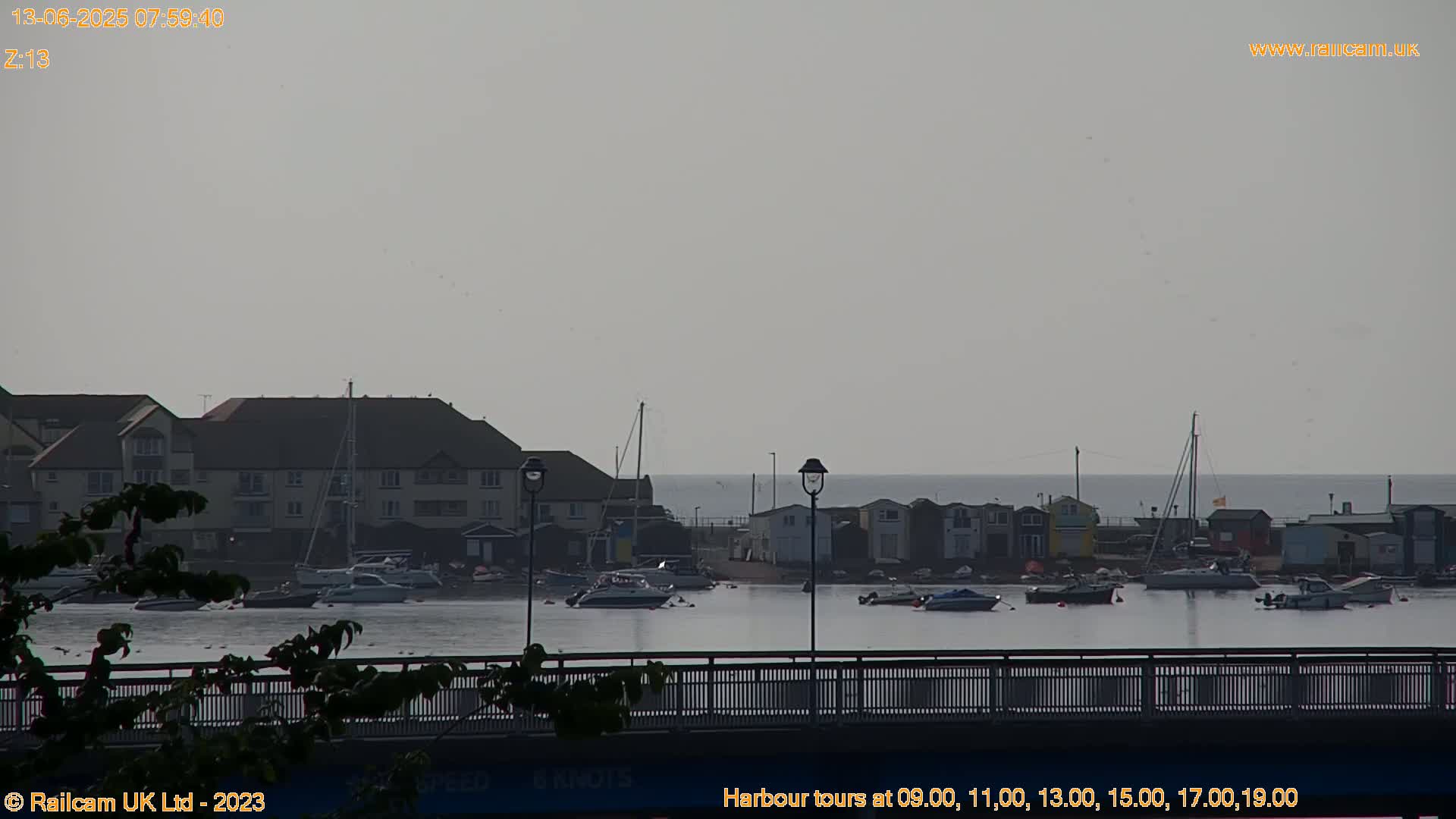 A calm, overcast day at a harbor shows several boats moored near a row of small buildings, viewed from across a bridge.
