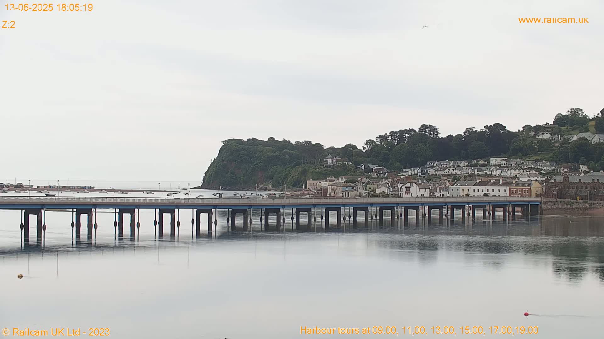 A long bridge spans a calm body of water, under an overcast sky, with a small town nestled on a hillside beyond.