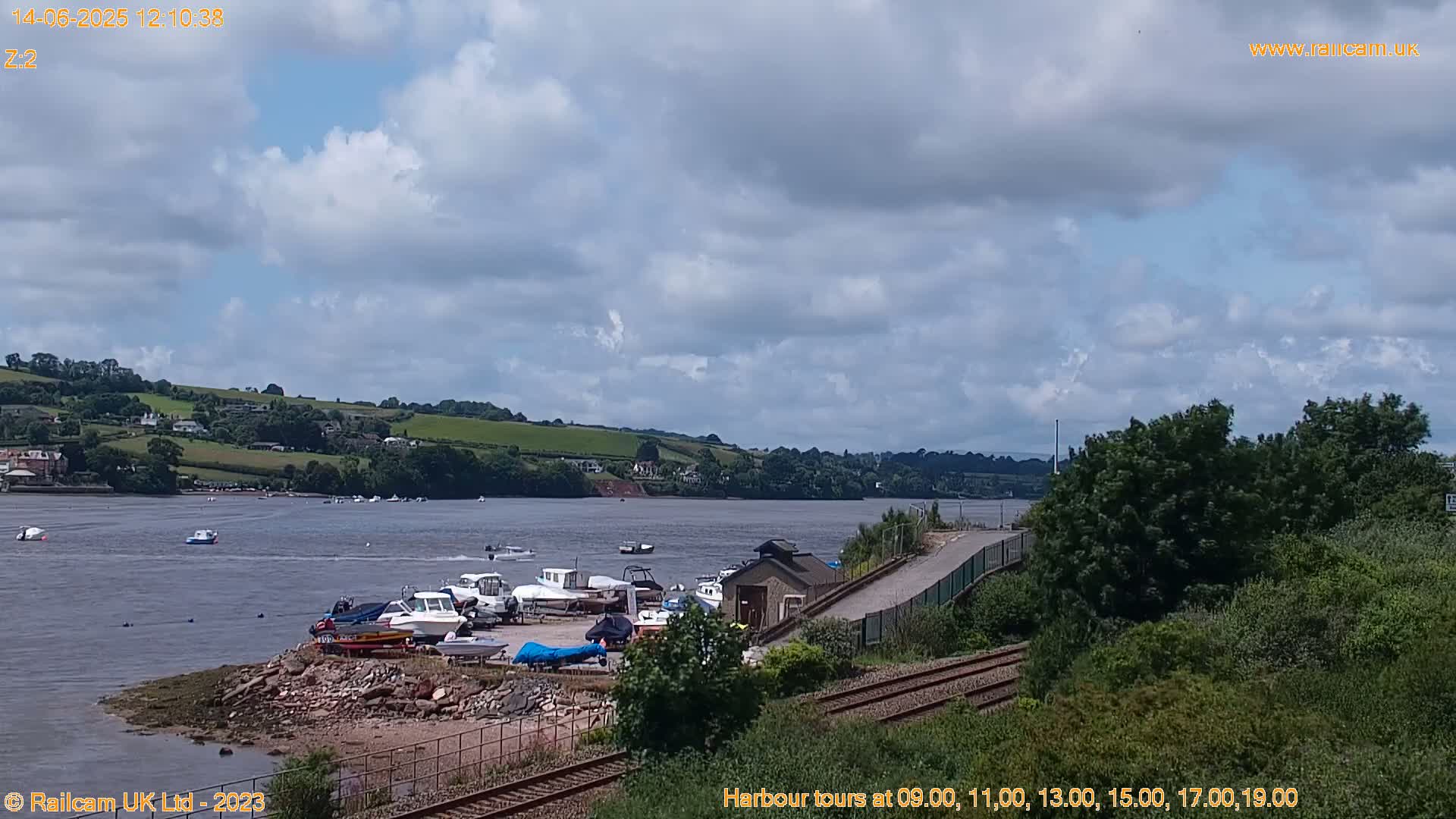 A partly cloudy day shows a river with several boats docked near a railway line and a small building; green hills are visible in the distance.