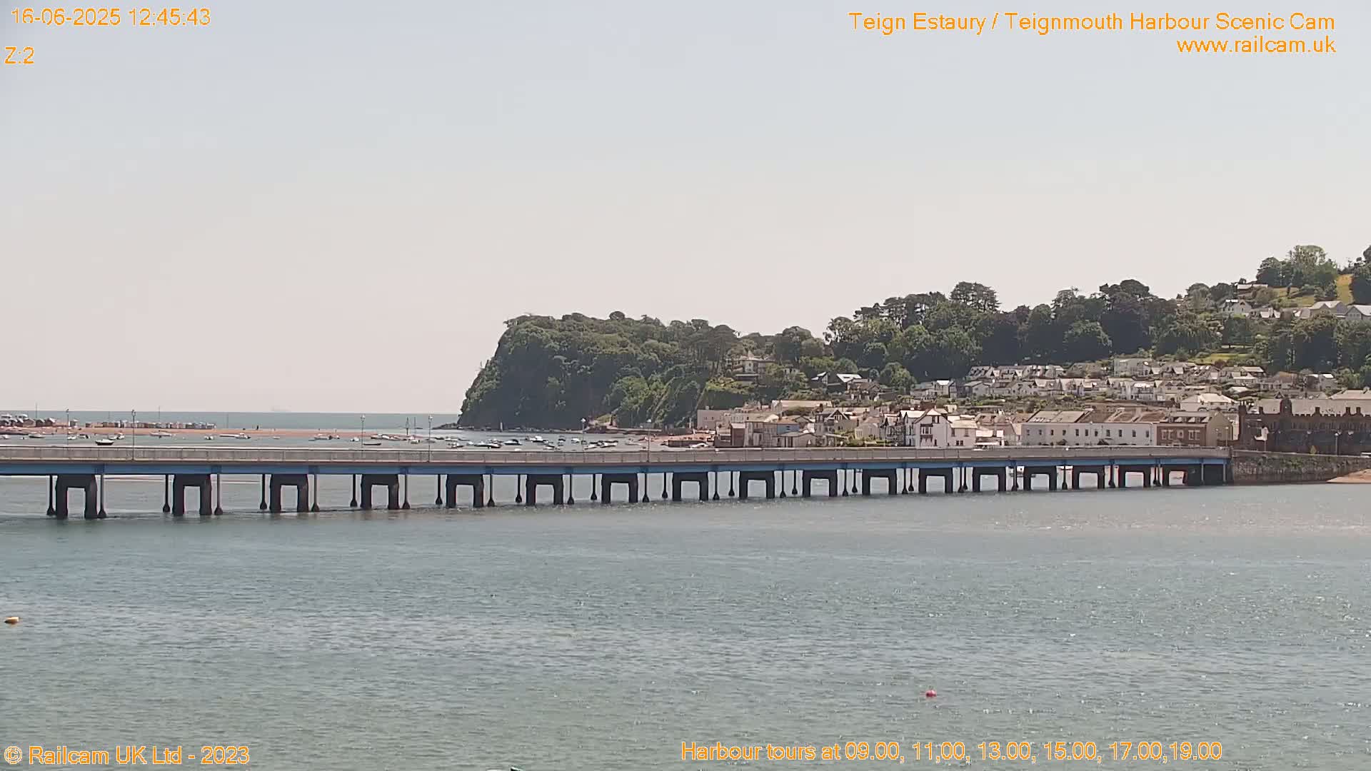 A long bridge stretches across calm water toward a town nestled against a verdant hillside under a bright, sunny sky.