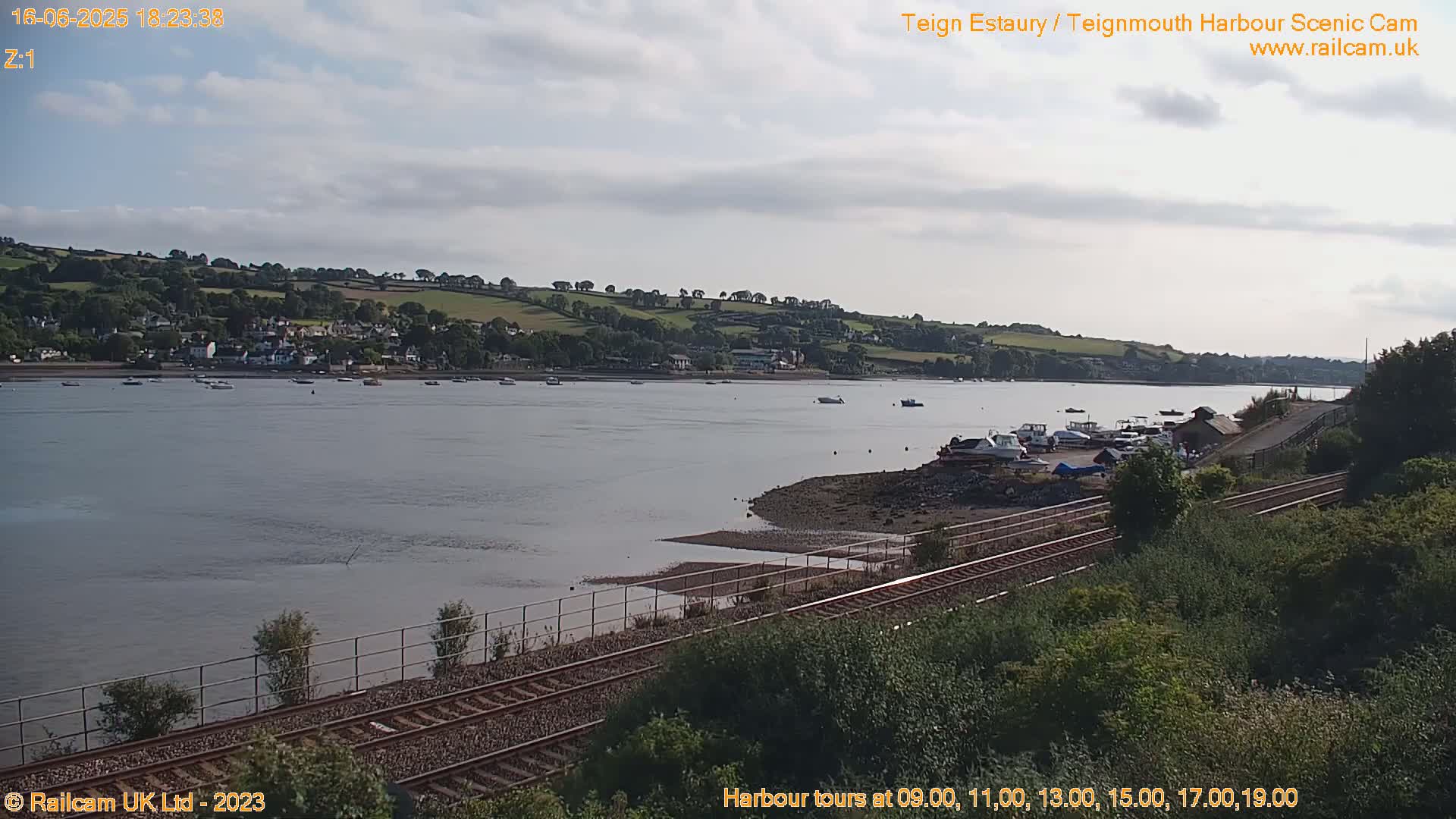 A calm river estuary with several boats moored near a shoreline with train tracks and lush green vegetation under a partly cloudy sky.