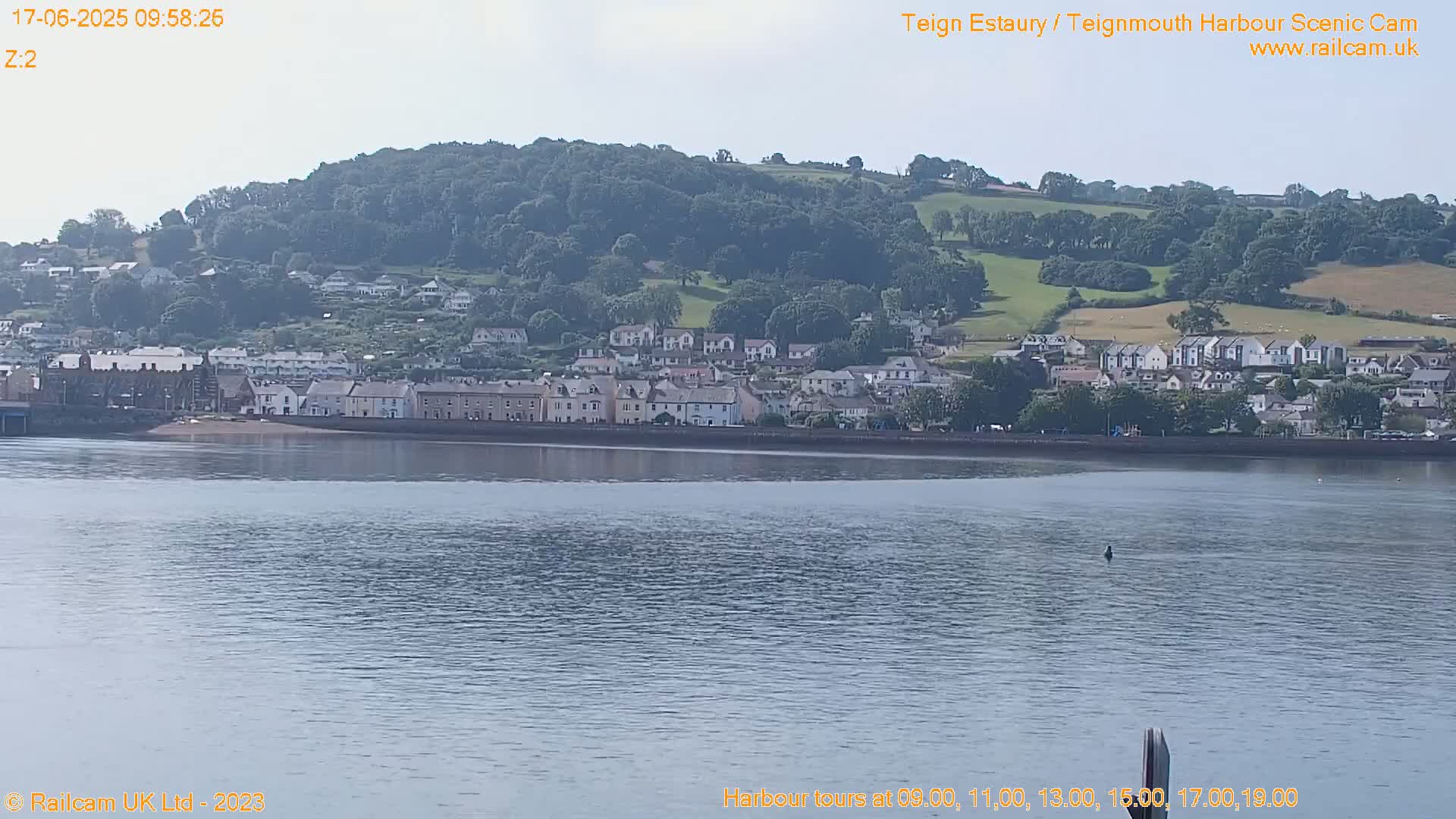 A seaside town sits on a calm body of water under a partly cloudy sky, backed by a verdant hill.