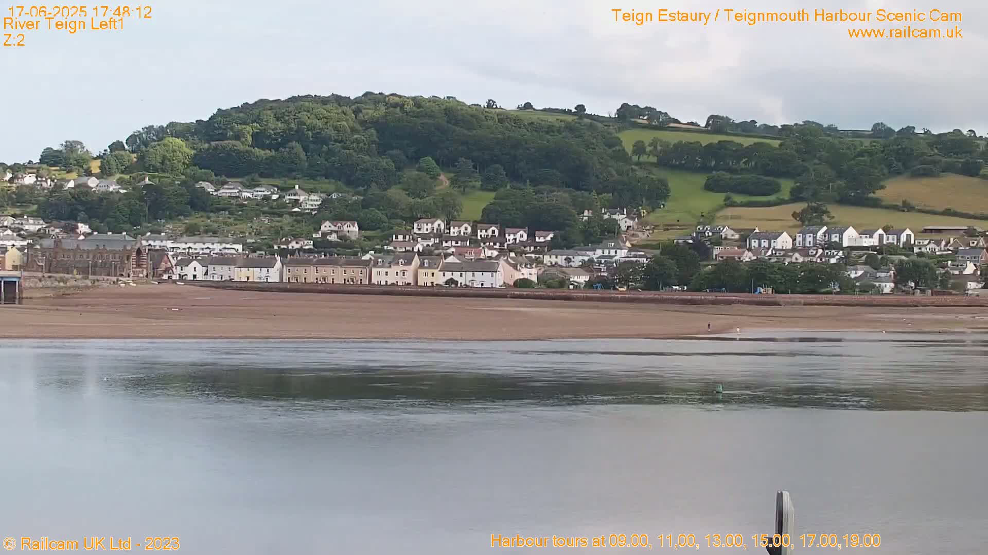 A coastal town nestled against a verdant hillside is visible across a calm body of water on a partly cloudy day.