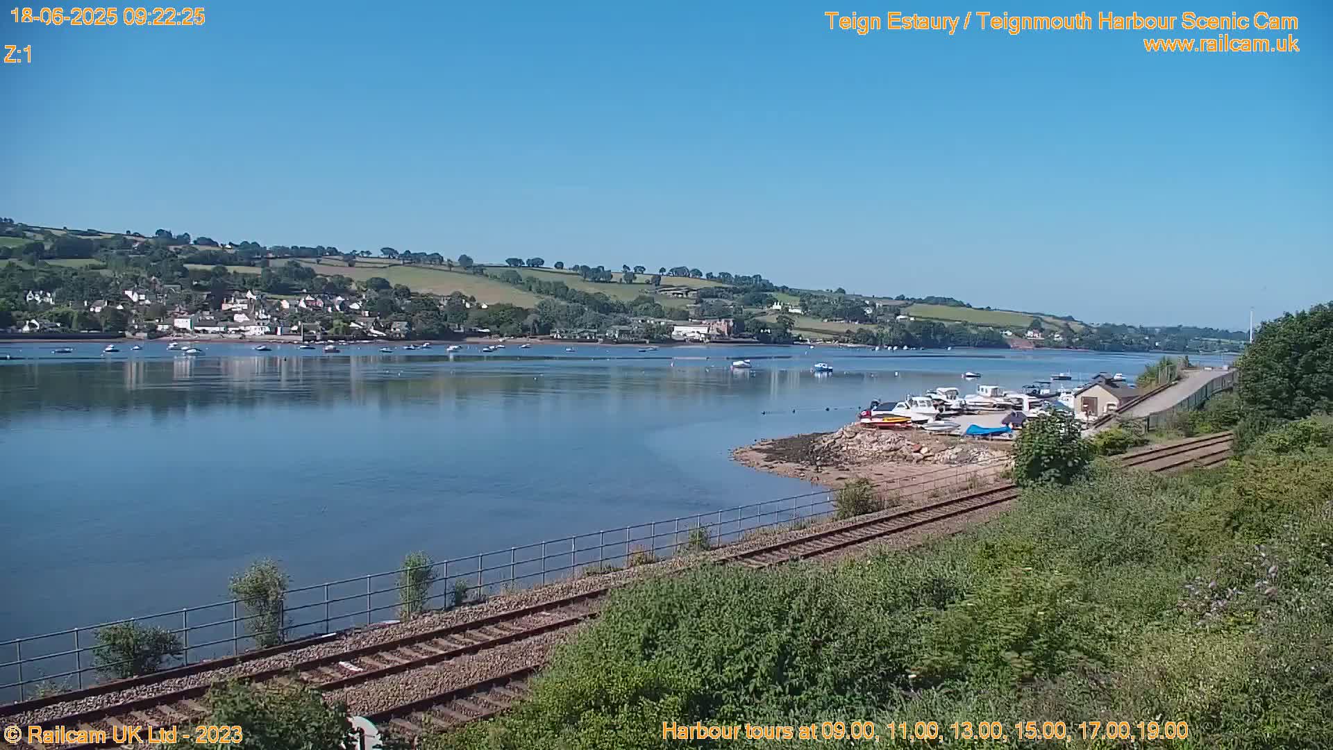 A calm, sunny day reveals a tranquil estuary scene with several boats moored near a shoreline, a railway line in the foreground, and a small town nestled on a hillside in the distance.