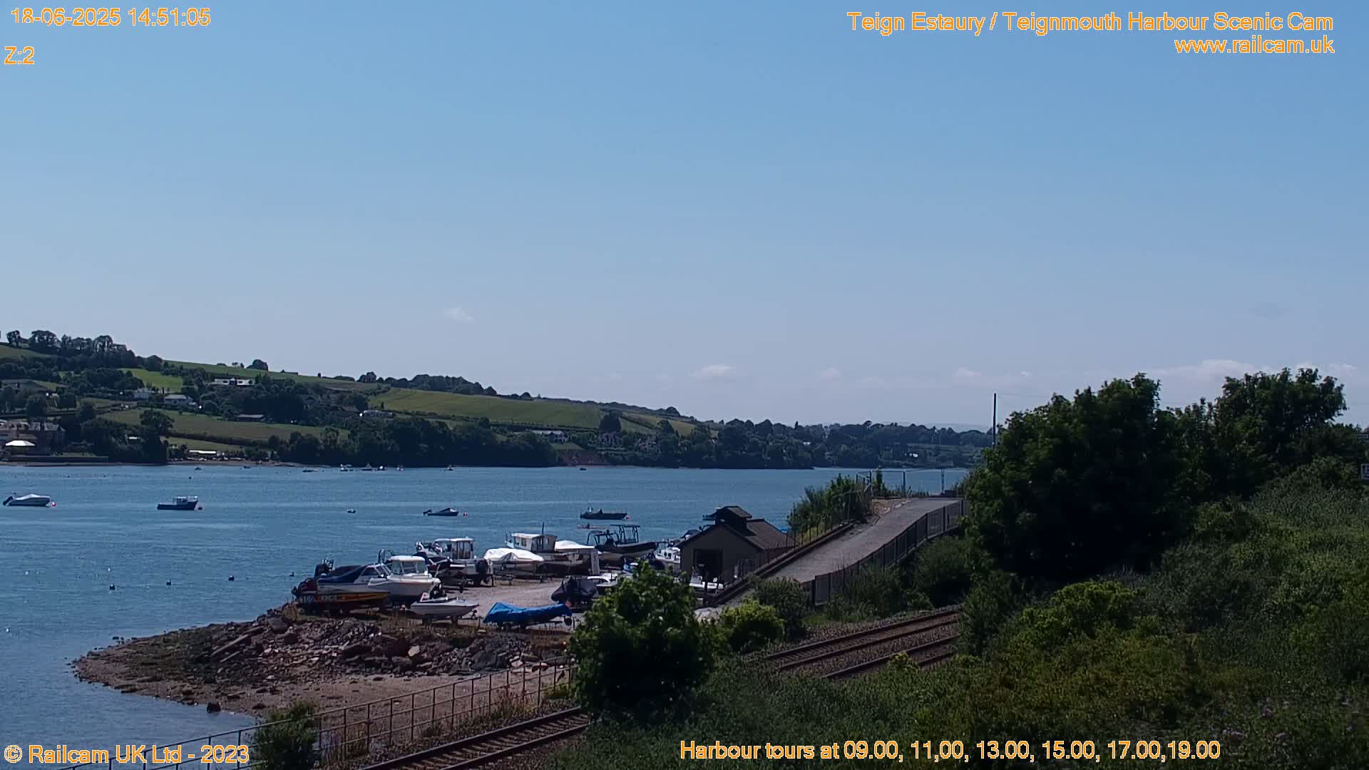 Several boats are docked at a harbor on a sunny day, with calm water and green hills visible in the distance.