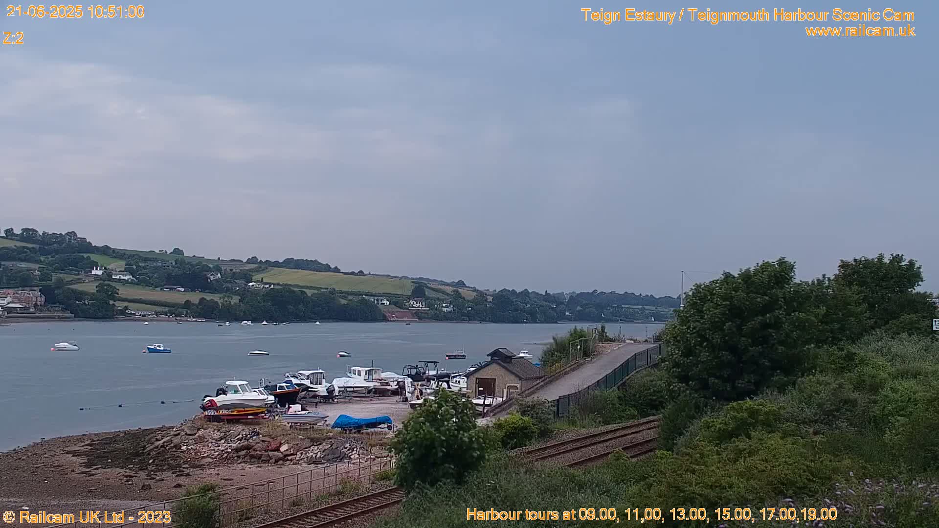 A calm, overcast day reveals a harbor scene with numerous boats docked near a small building, situated next to a railway track and lush green hillsides overlooking a body of water.