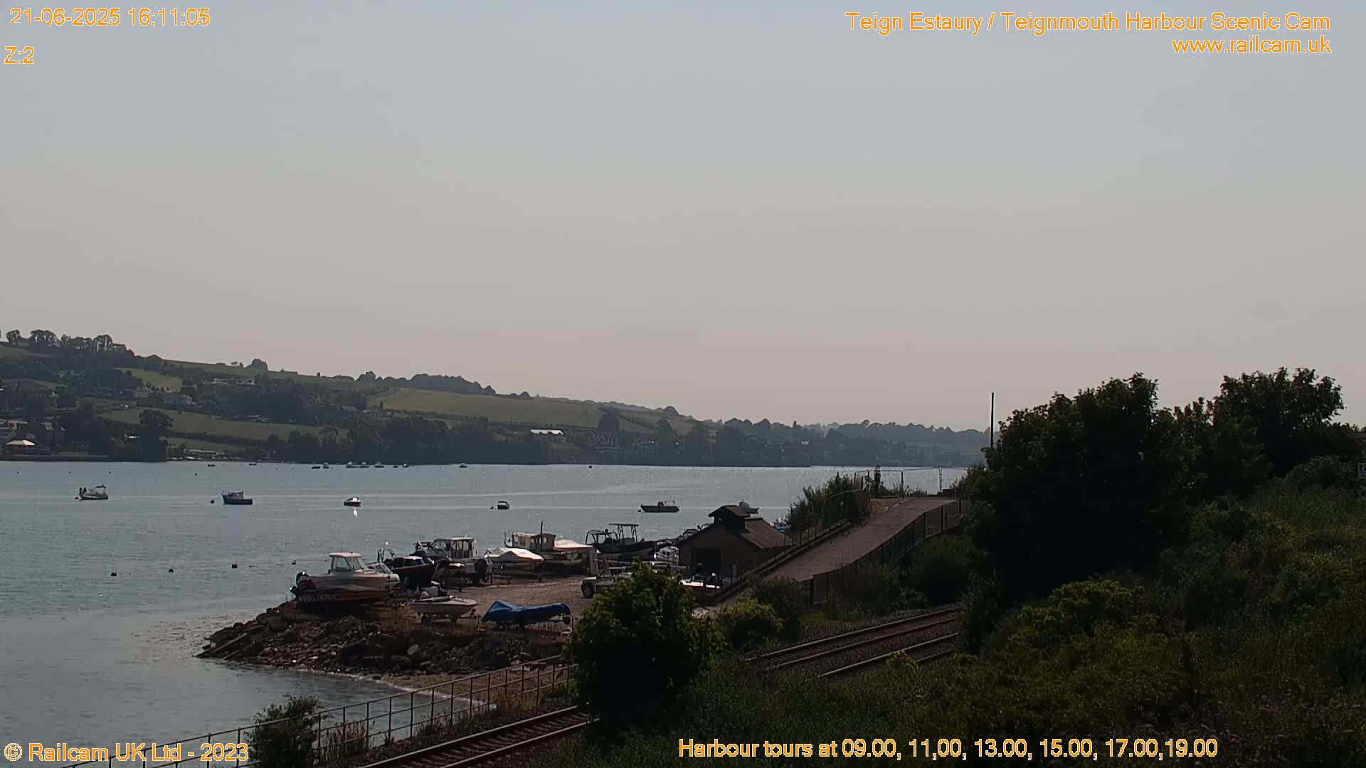 Several boats are moored near a harbor building, next to train tracks, with calm water and a hazy sky in the background.