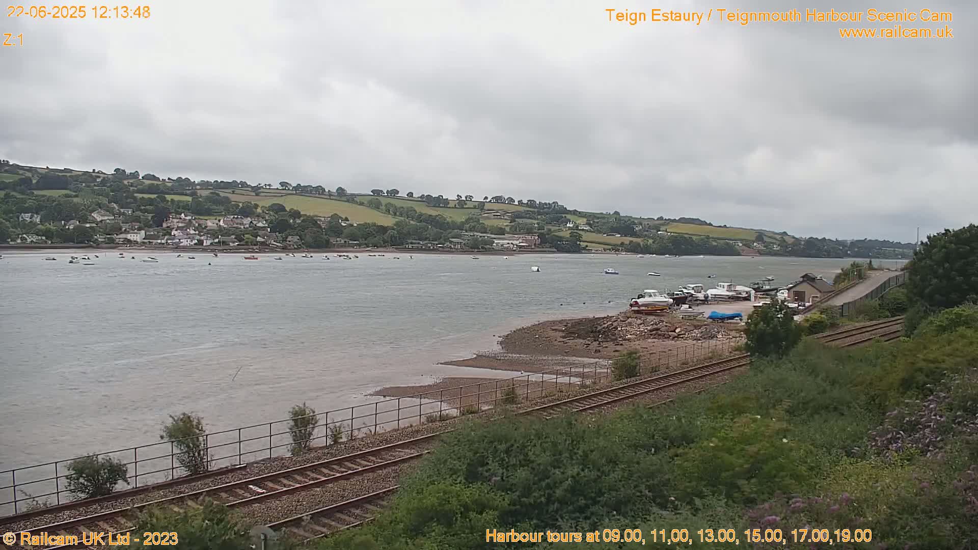 A wide shot of a calm river with numerous boats, a small town on the far bank, and train tracks in the foreground under an overcast sky.