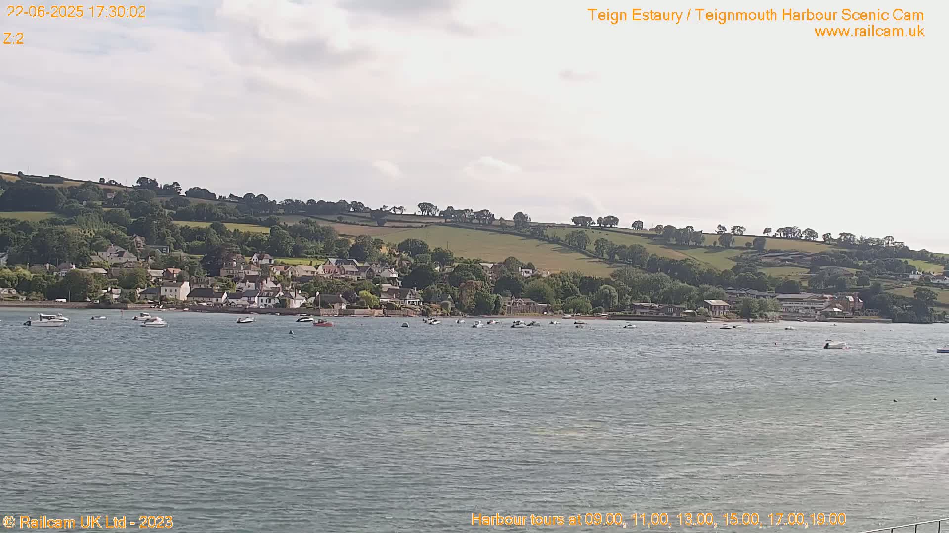A calm body of water shows several small boats near a shoreline village nestled at the base of a gently sloping green hill under a partly cloudy sky.