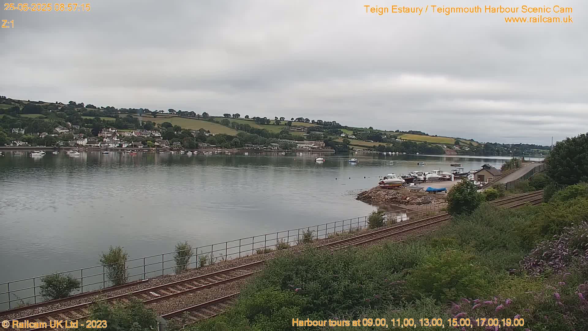 A calm body of water stretches towards a hillside town under an overcast sky, with railroad tracks and docked boats in the foreground.
