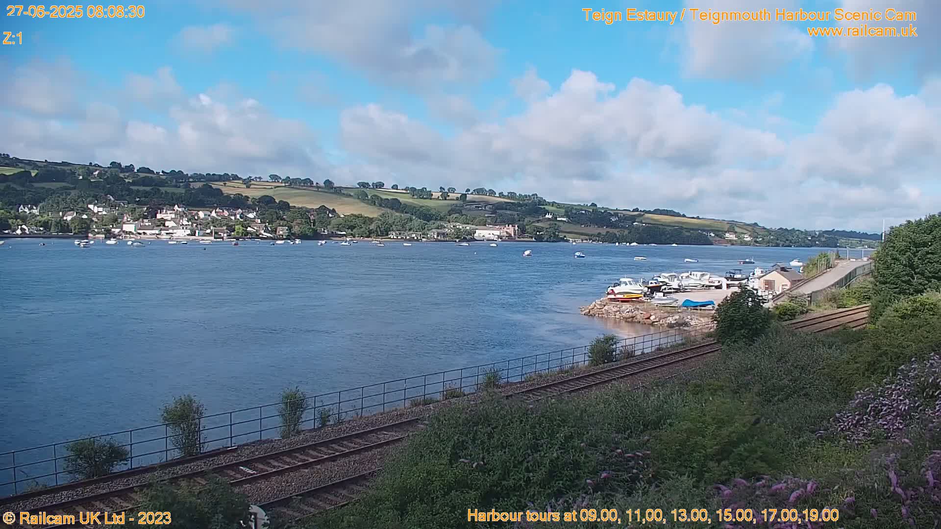 A calm body of water with numerous boats, a town nestled on a hillside, and railroad tracks in the foreground under a partly cloudy sky.