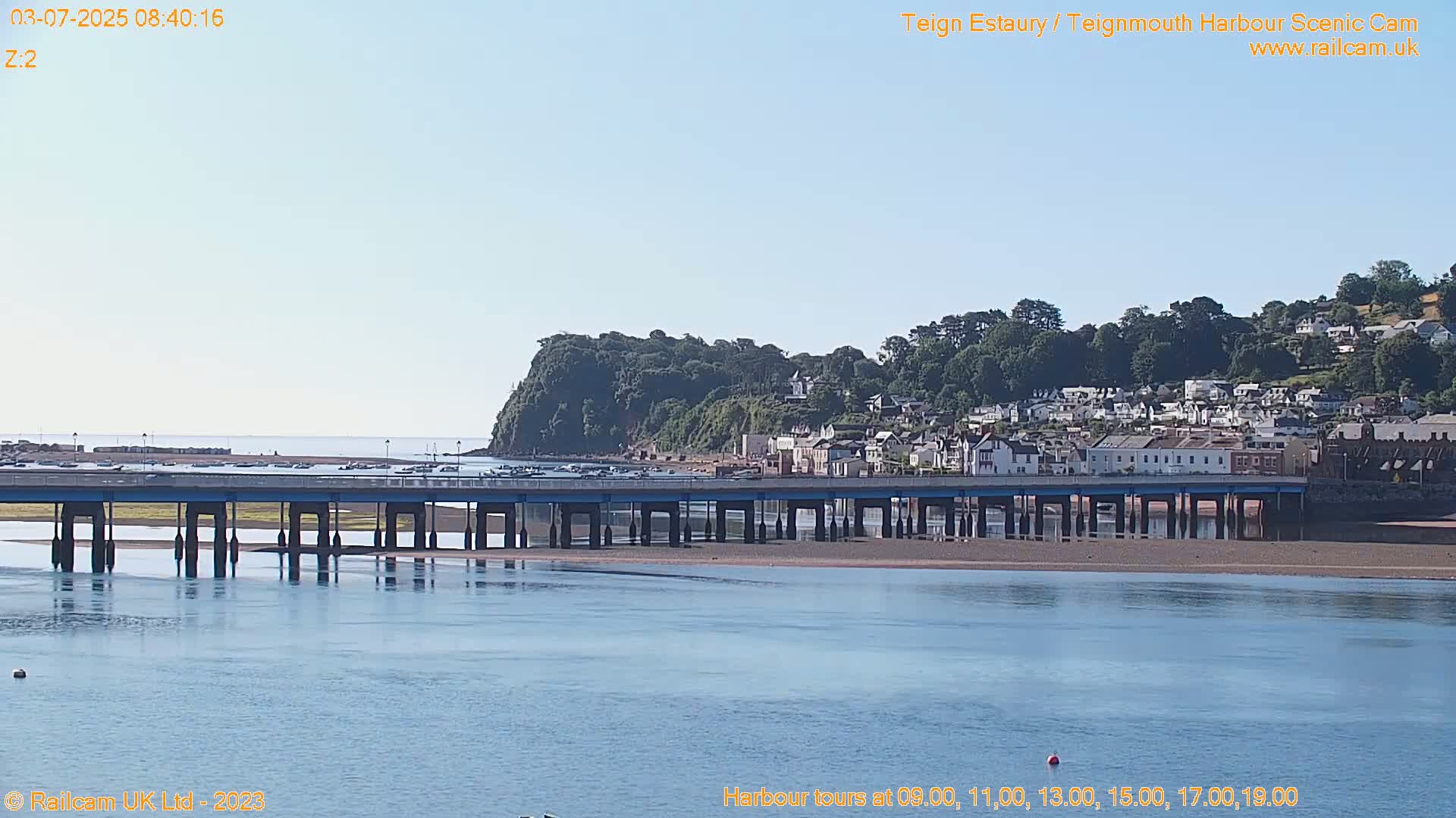A long bridge stretches across a calm body of water under a clear, bright sky, with a town and a hill covered in lush greenery visible in the background.