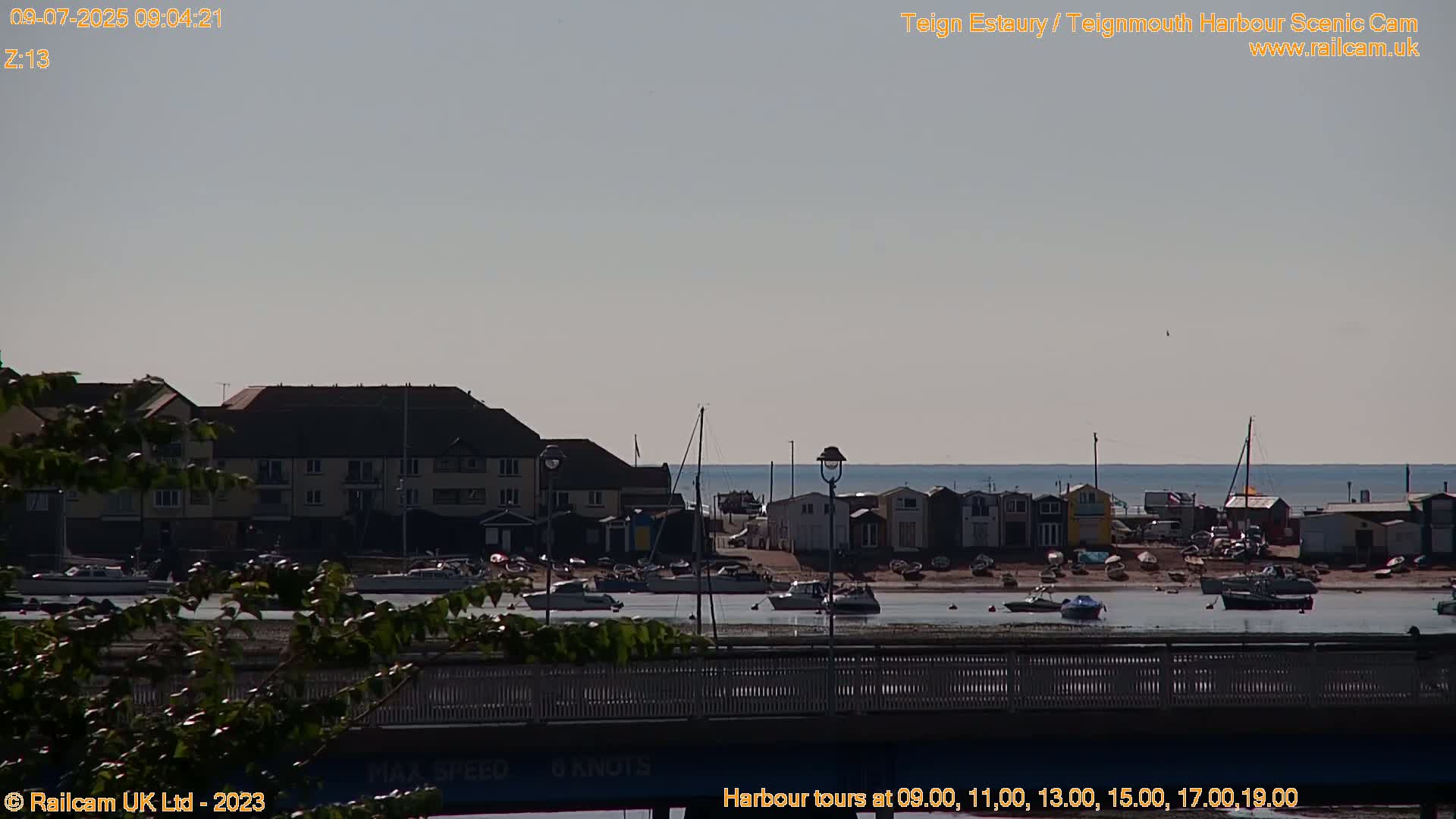 A calm, sunny day shows a harbor with boats, buildings, and a beach, viewed from a bridge.