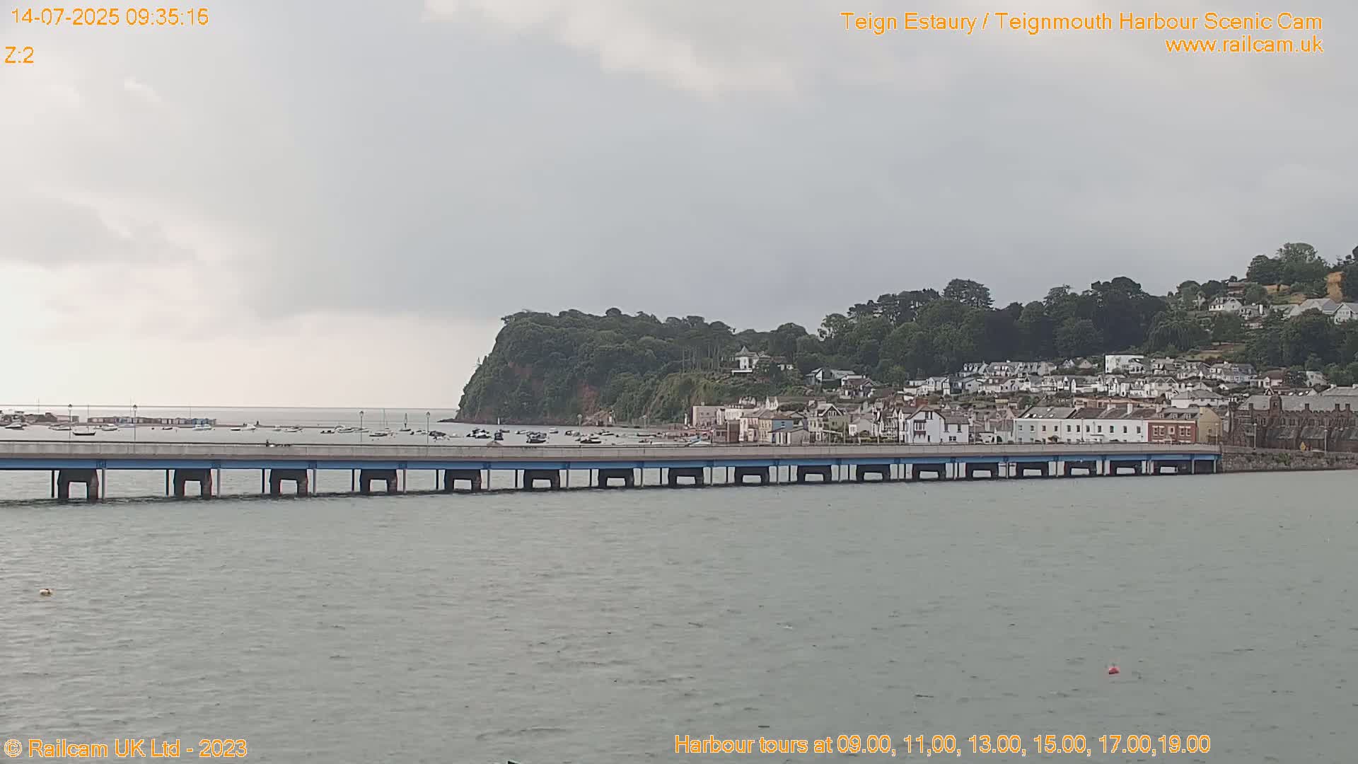 A long bridge stretches across a calm body of water toward a town nestled on a hillside under an overcast sky.