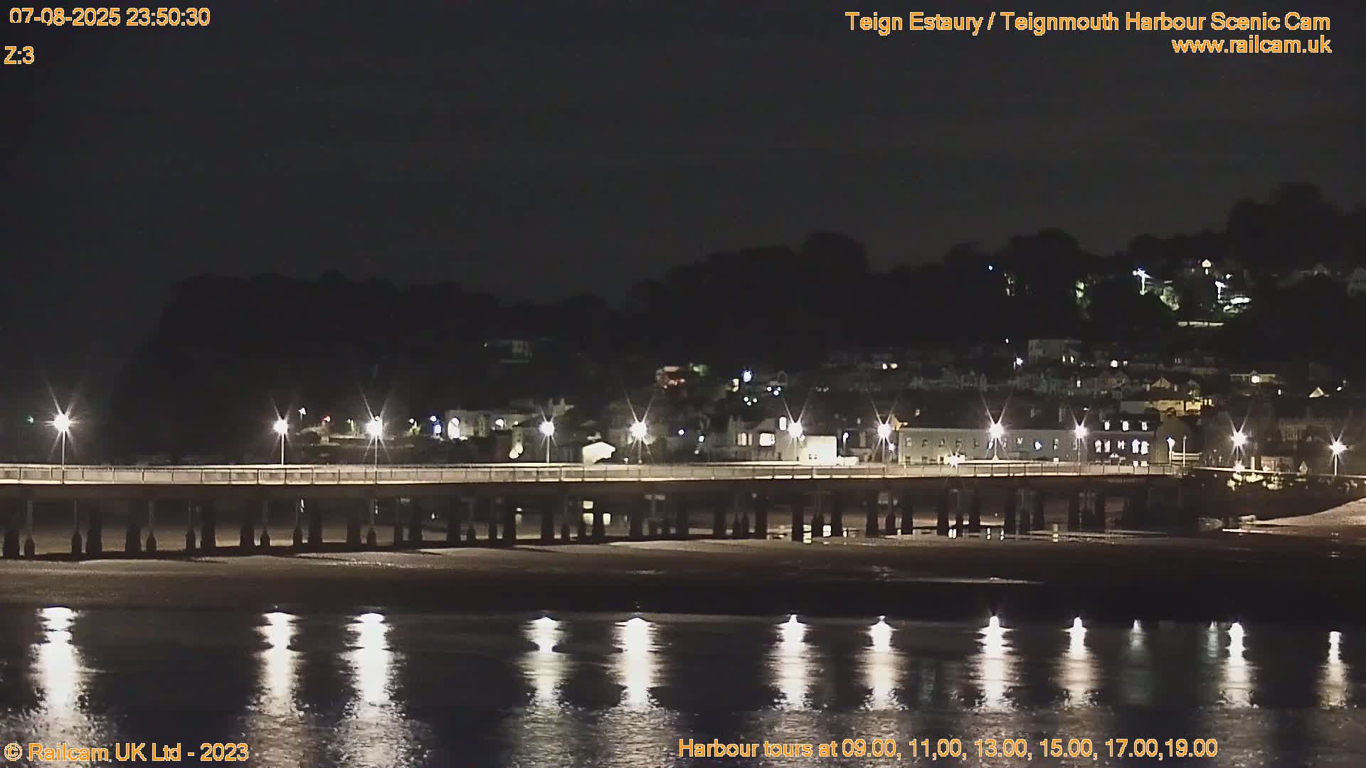 A long pier lit by lamps stretches across a calm, dark body of water at night, with a hillside town visible in the background.