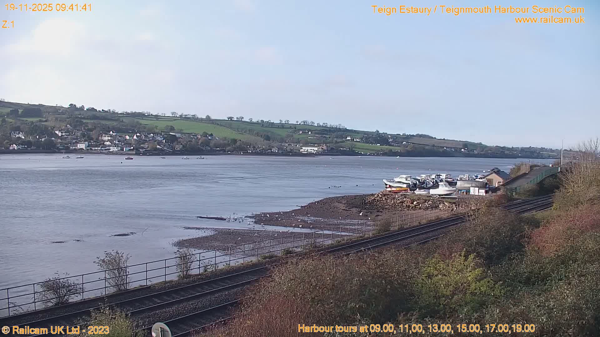 A hazy, bright outdoor scene overlooks a wide body of water with a pier and ducks in the foreground, leading to waterfront buildings nestled against tree-covered hills under hazy sunshine.