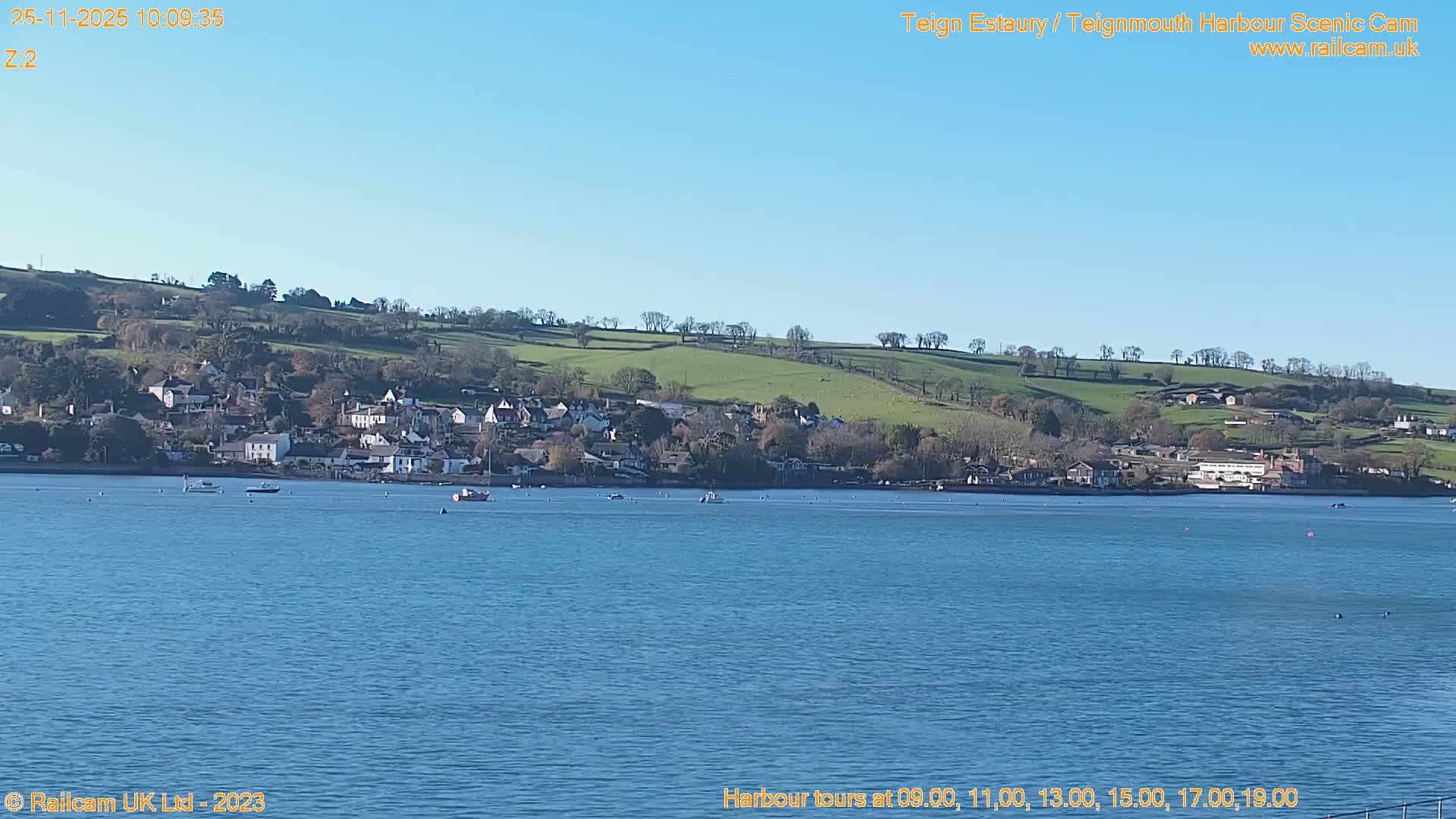 A bright sunny day illuminates a wide blue estuary dotted with several boats, a charming town with white buildings along its distant shore, and verdant rolling hills in the background.