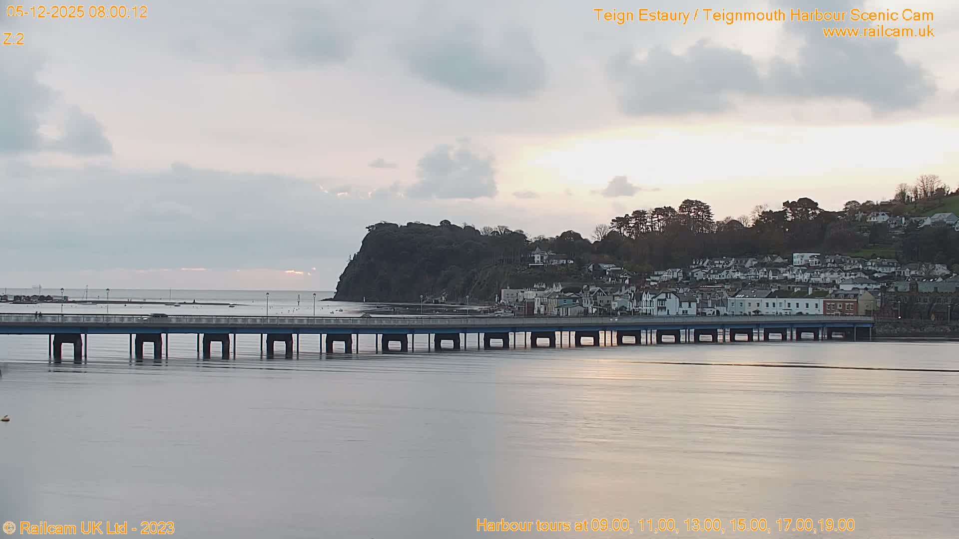 A long pier stretches across calm estuary waters towards a hillside town backed by a tree-covered headland, all under an overcast sky with a hint of golden light on the morning horizon.