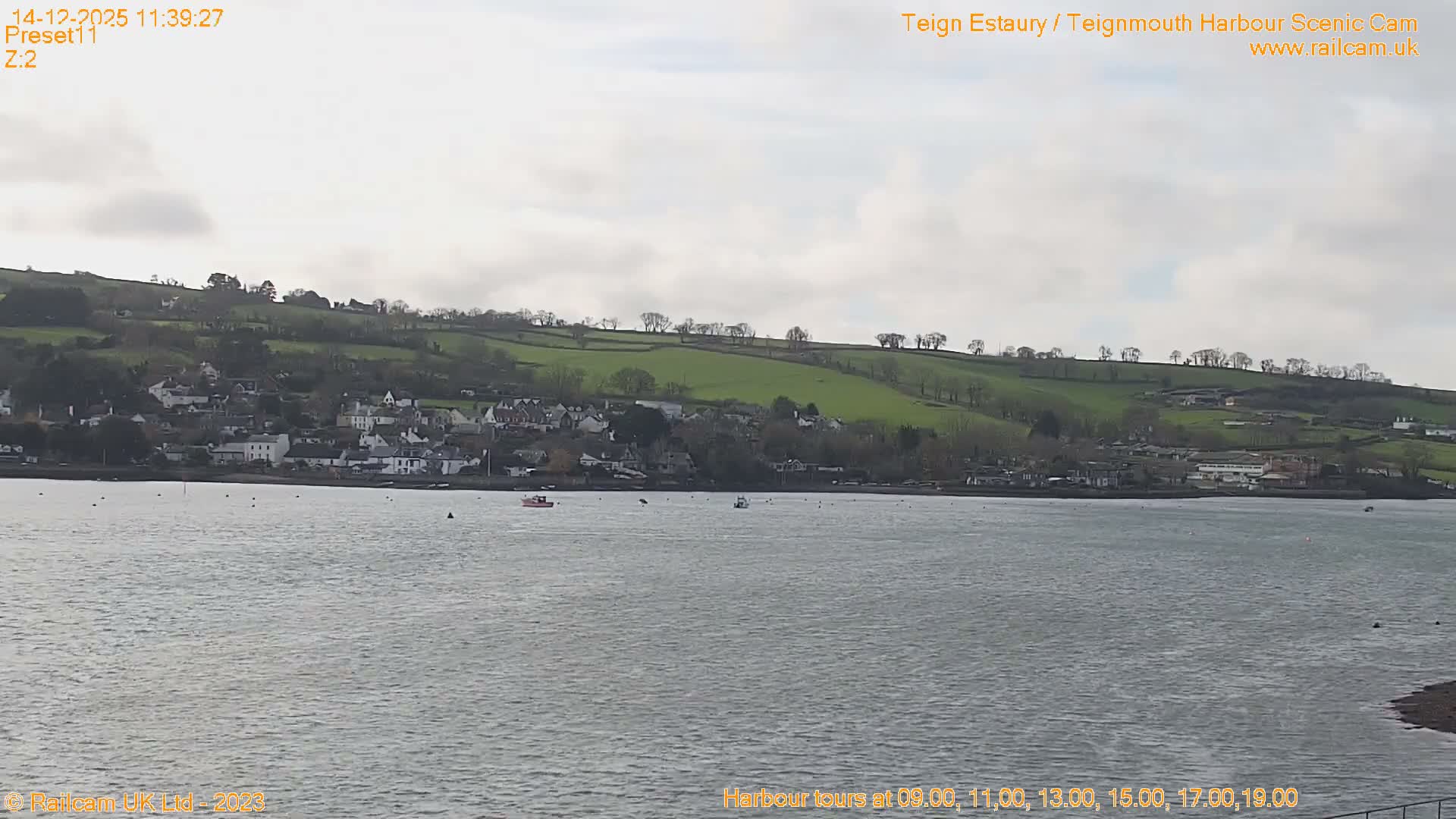 A long pier stretches across calm estuary waters towards a hillside town backed by a tree-covered headland, all under an overcast sky with a hint of golden light on the morning horizon.