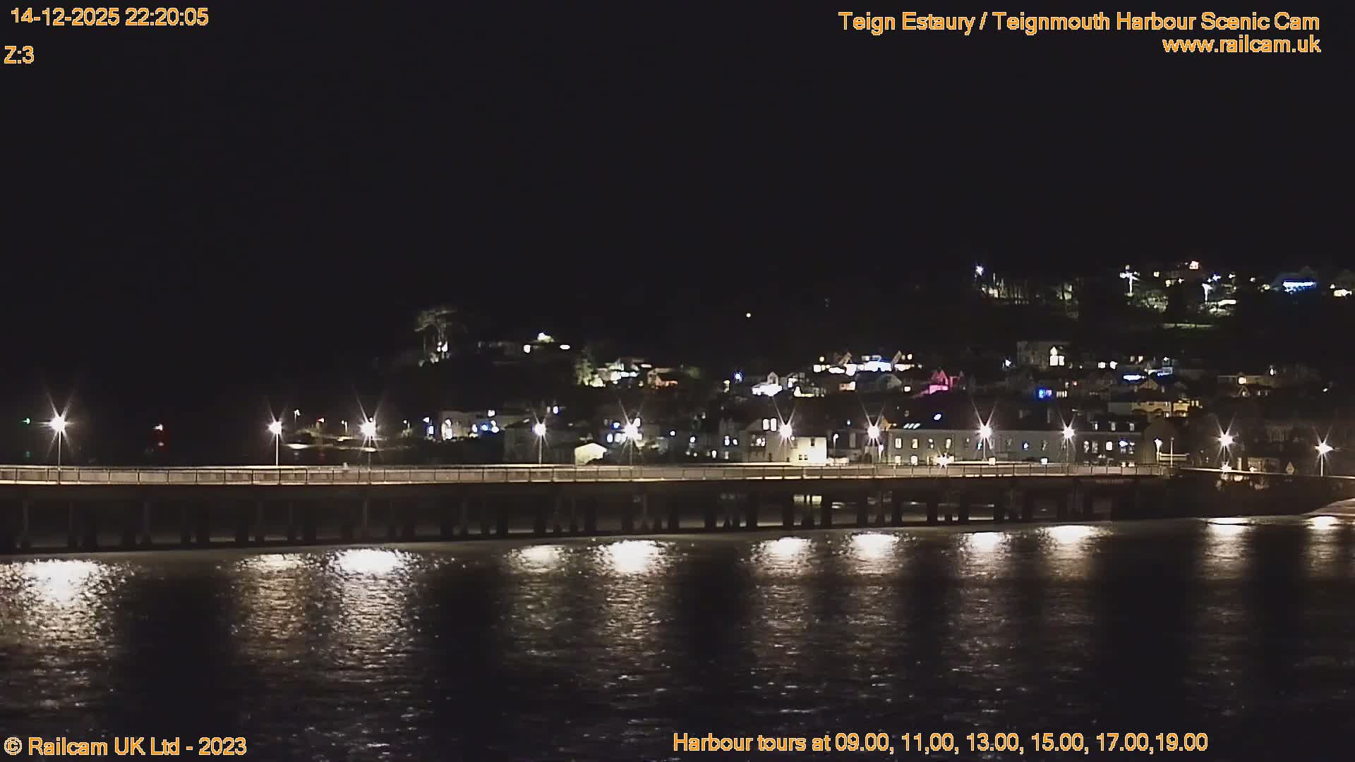 A long pier stretches across calm estuary waters towards a hillside town backed by a tree-covered headland, all under an overcast sky with a hint of golden light on the morning horizon.