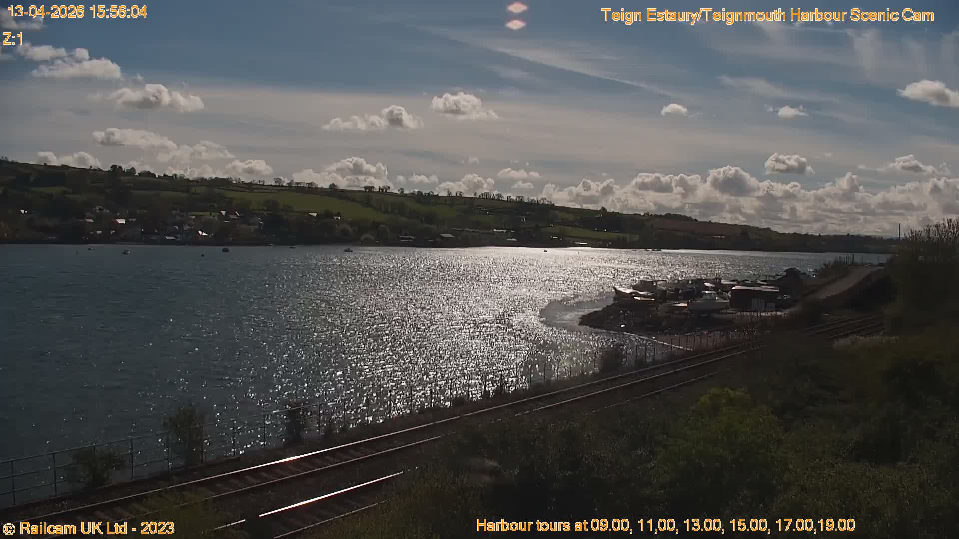A long pier stretches across calm estuary waters towards a hillside town backed by a tree-covered headland, all under an overcast sky with a hint of golden light on the morning horizon.