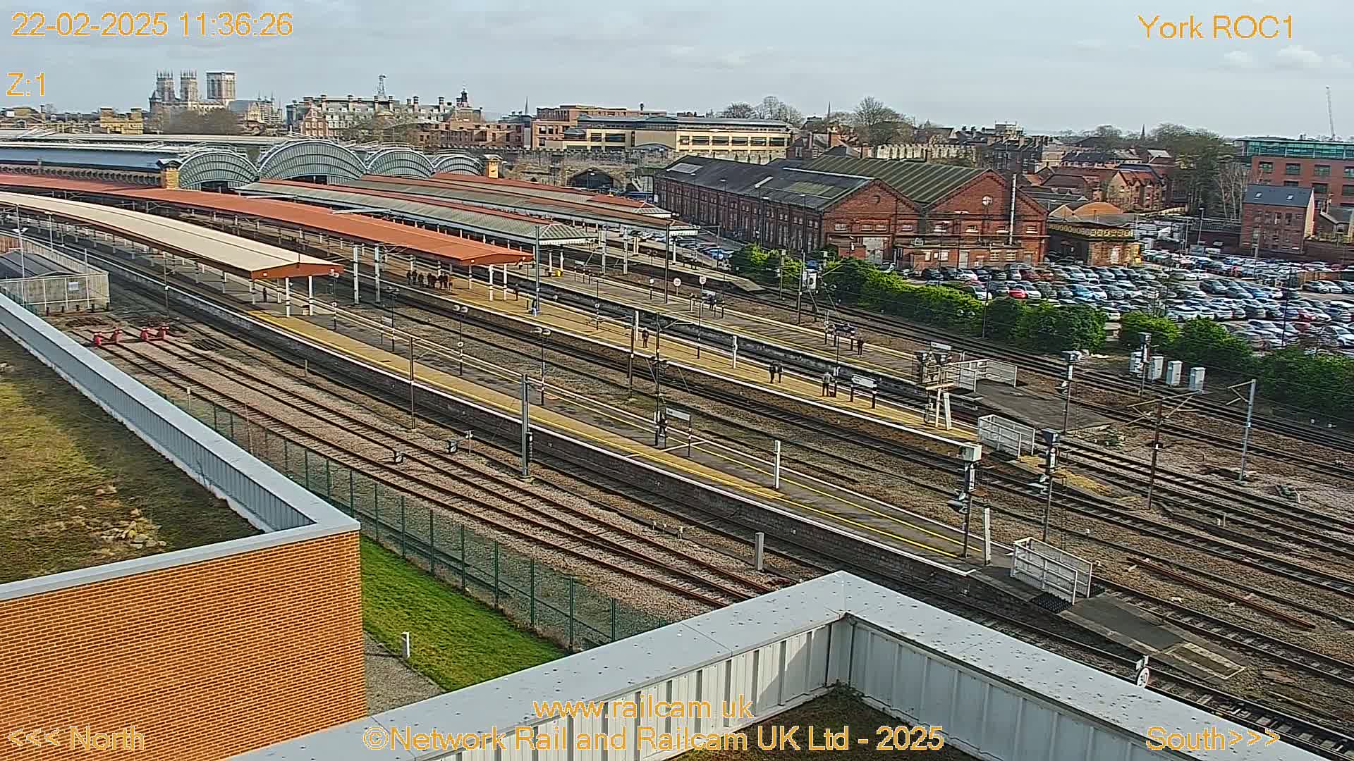 A high-angle view of a train station on a sunny day, showing numerous tracks, platforms, and a large parking lot, with a city skyline visible in the background.