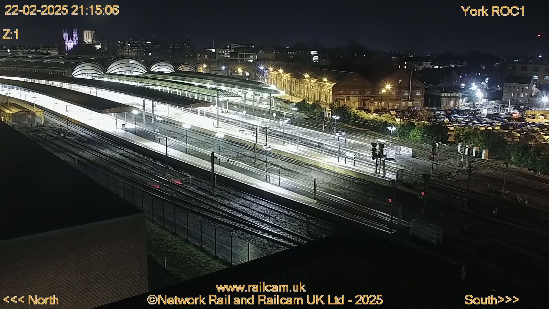 A brightly lit train station at night with many tracks and platforms, surrounded by buildings and a large parking lot.
